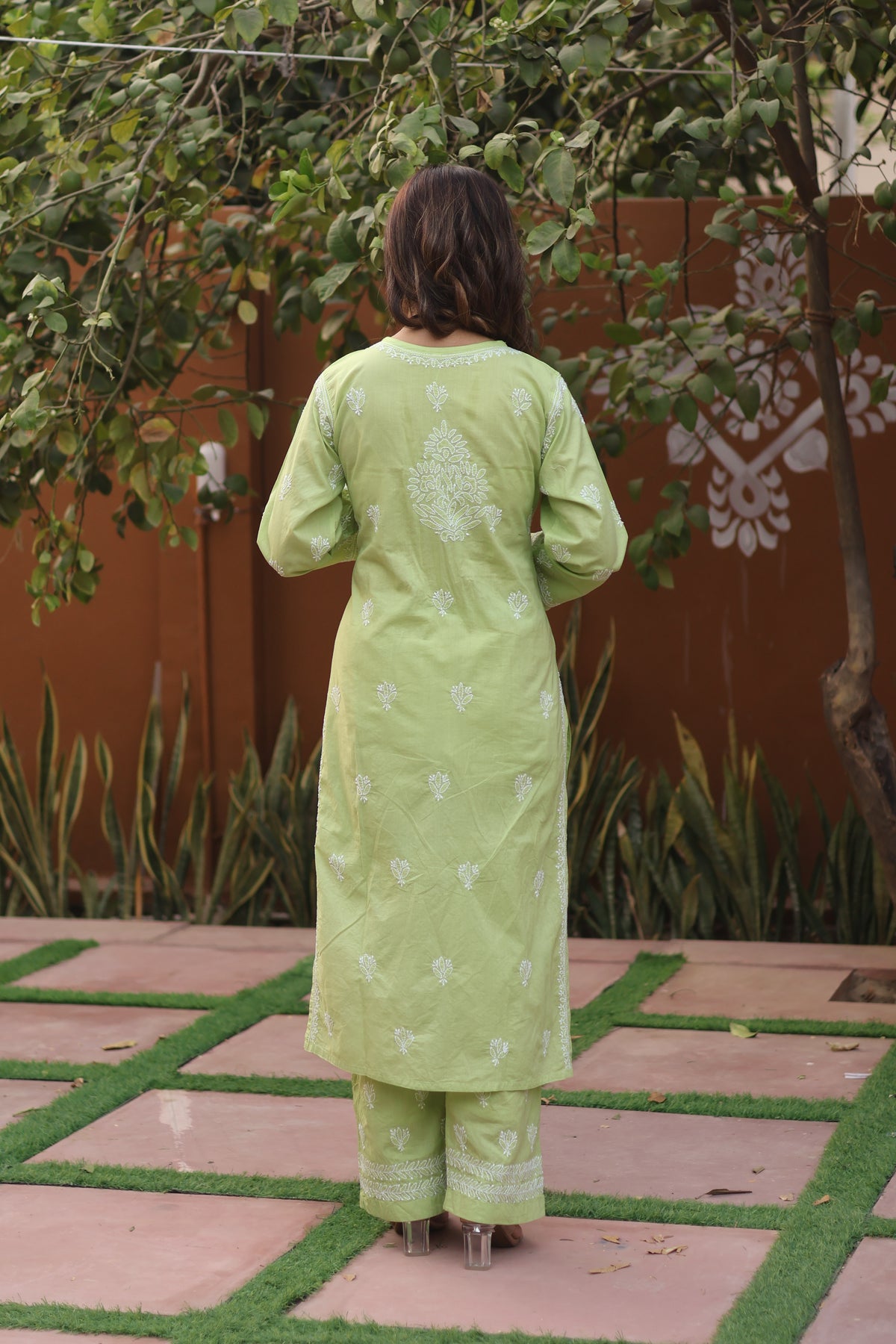 A woman viewed from behind wearing a light green traditional kurta and trouser set with white embroidery, standing on a tiled outdoor patio with plants and a terracotta wall in the background.