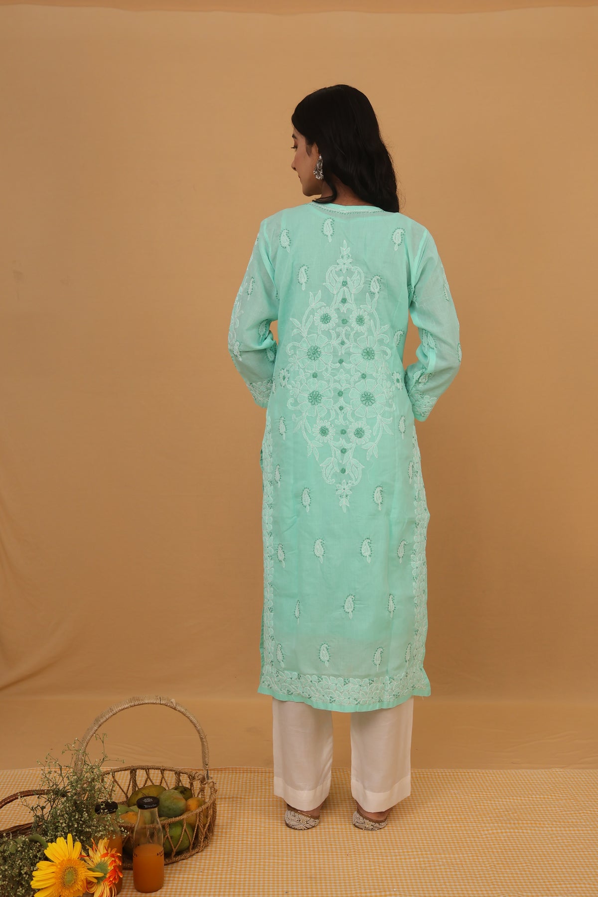 A woman stands with her back to the camera wearing a mint green embroidered kurta and white pants against a solid beige backdrop.