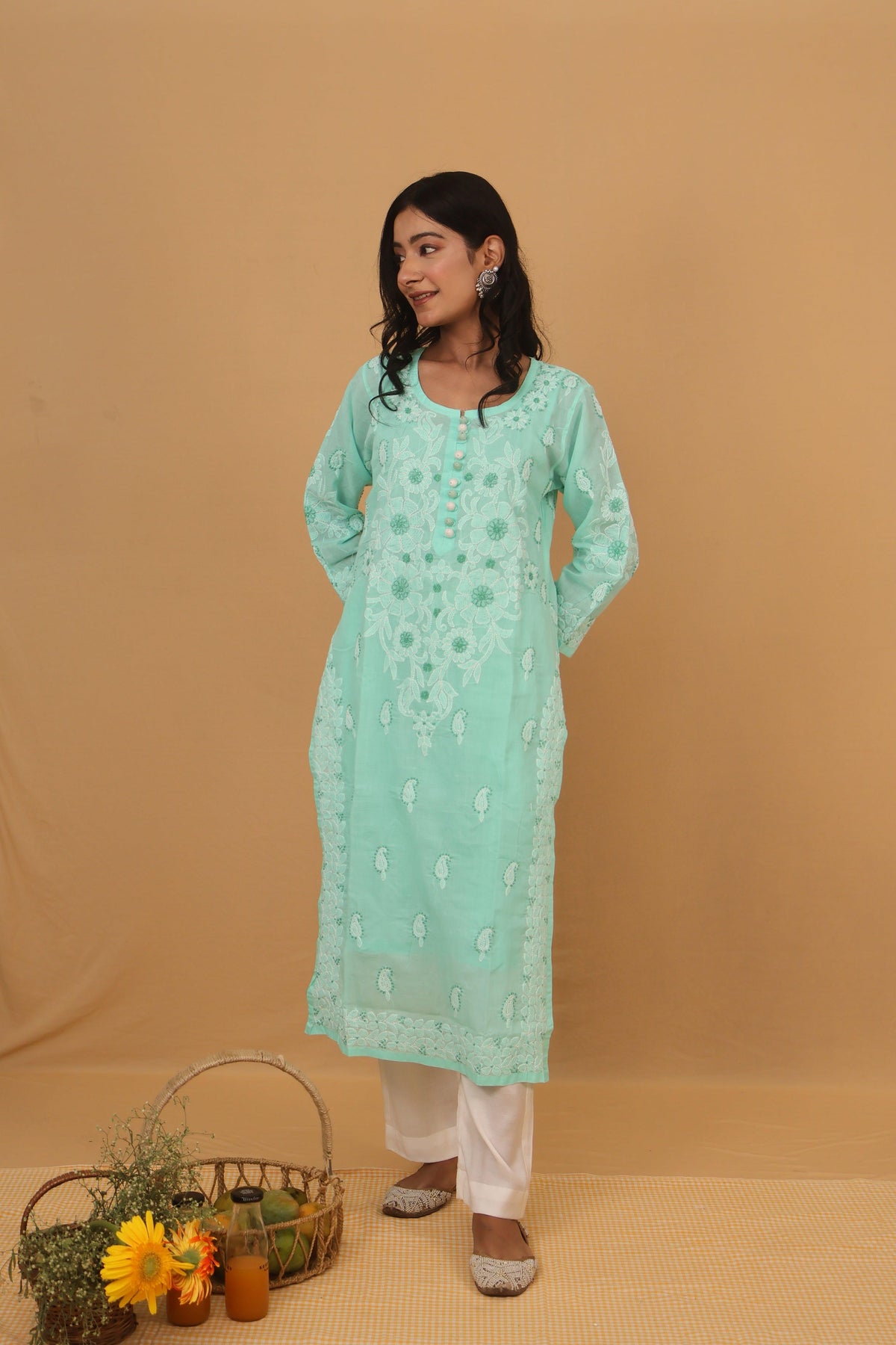 A woman modeling a mint green embroidered kurta with white pants, standing against a solid beige backdrop. A small decorative basket with flowers is on the floor to the left.