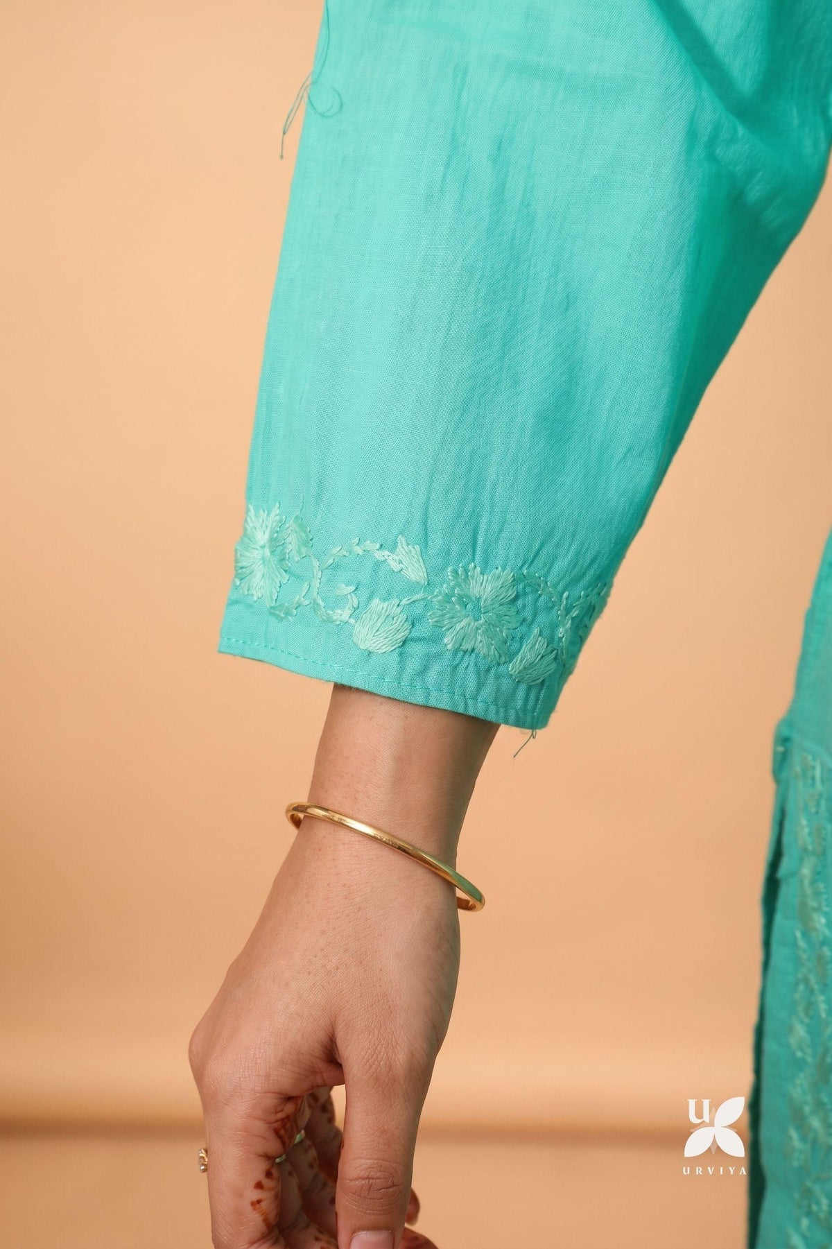 Close-up of a person's hand and forearm wearing a turquoise sleeve with floral lace trim, against a warm peach background. A thin gold bangle is visible on the wrist.
