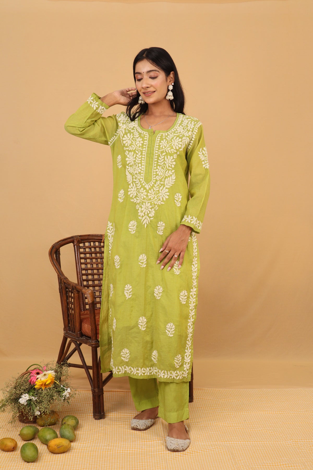 A woman models a light green three-piece salwar kameez suit with white embroidery, standing next to a brown wooden chair against a solid beige backdrop.