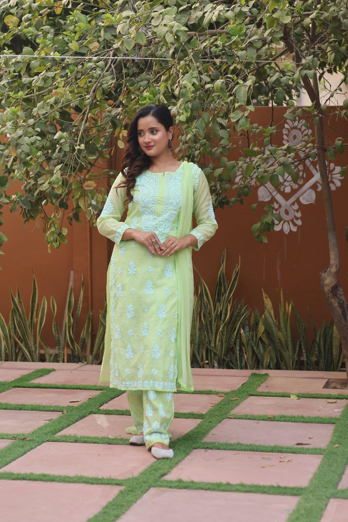 A woman wearing a light green ethnic salwar kameez suit with white embroidery, standing outdoors on a tiled path with green plants and a terracotta-colored wall in the background. Green Mul Chanderi White Thread Chikankari Ghaspatti 3 Piece Set with Dupatta