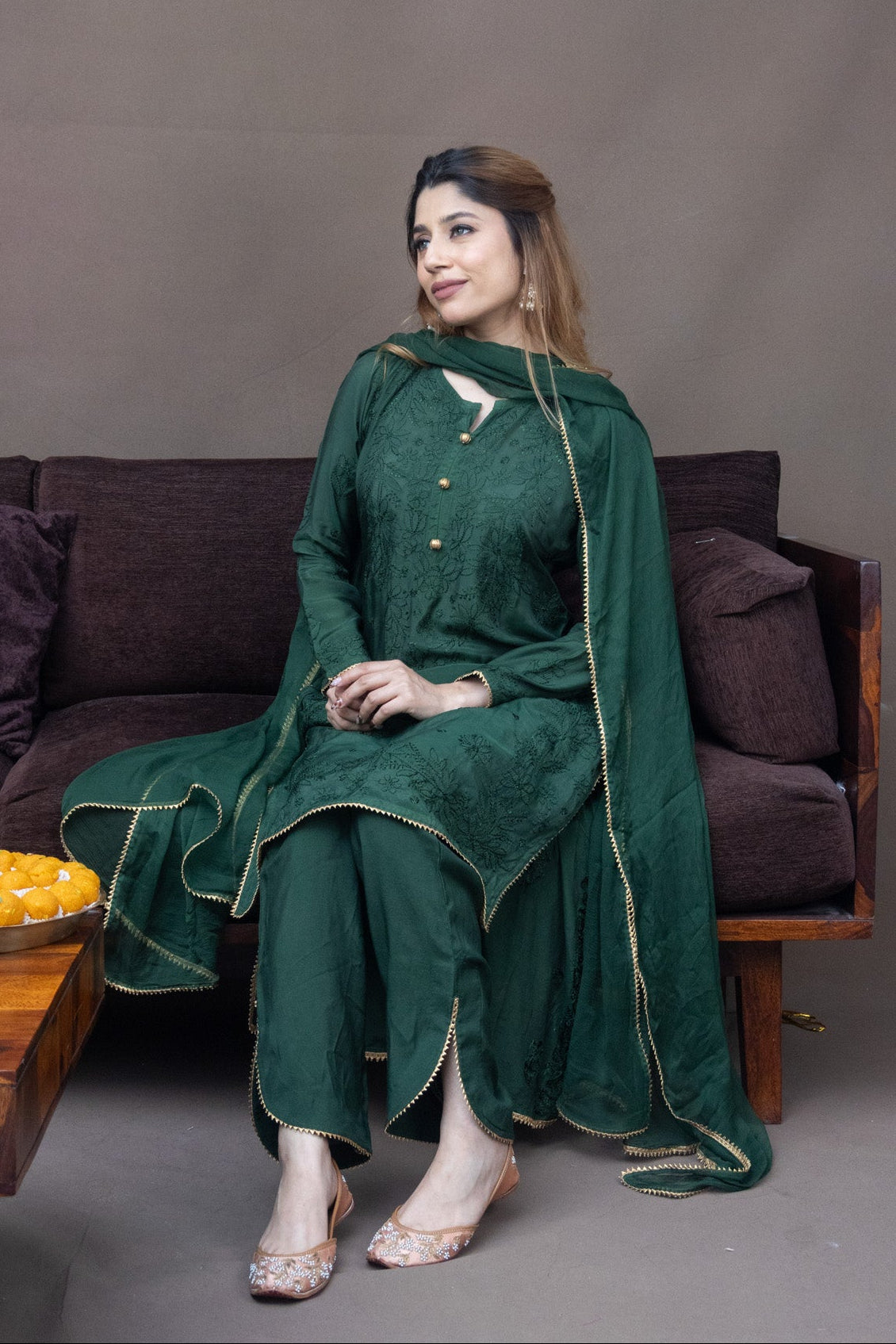 A woman wearing a dark green traditional outfit with gold trim sits on a dark brown sofa. The outfit includes a long-sleeved kurta with gold buttons and a matching draped shawl.