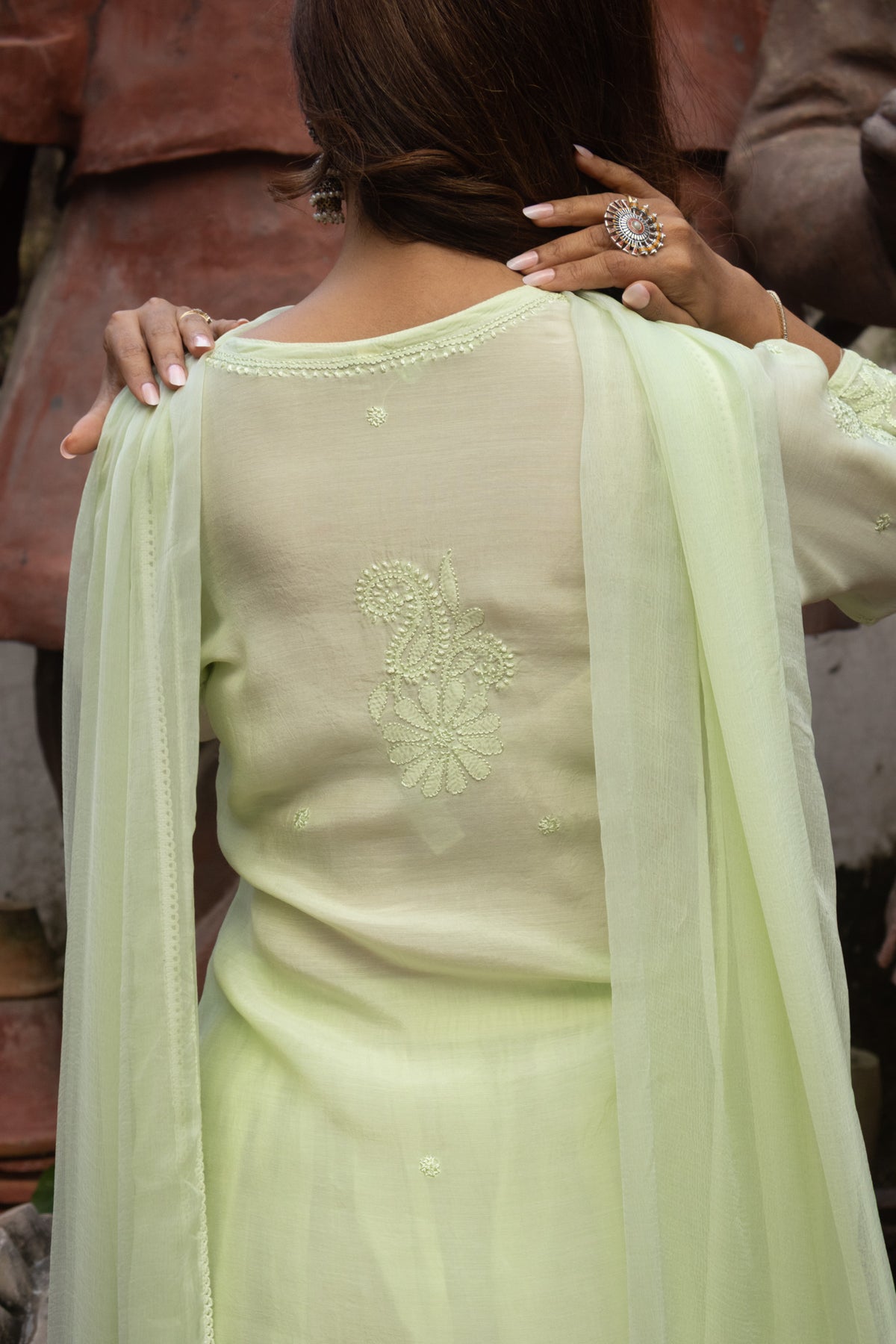 A woman shown from the back wearing a light pastel green traditional outfit with embroidered detailing on the back and a matching sheer dupatta draped over her shoulders.