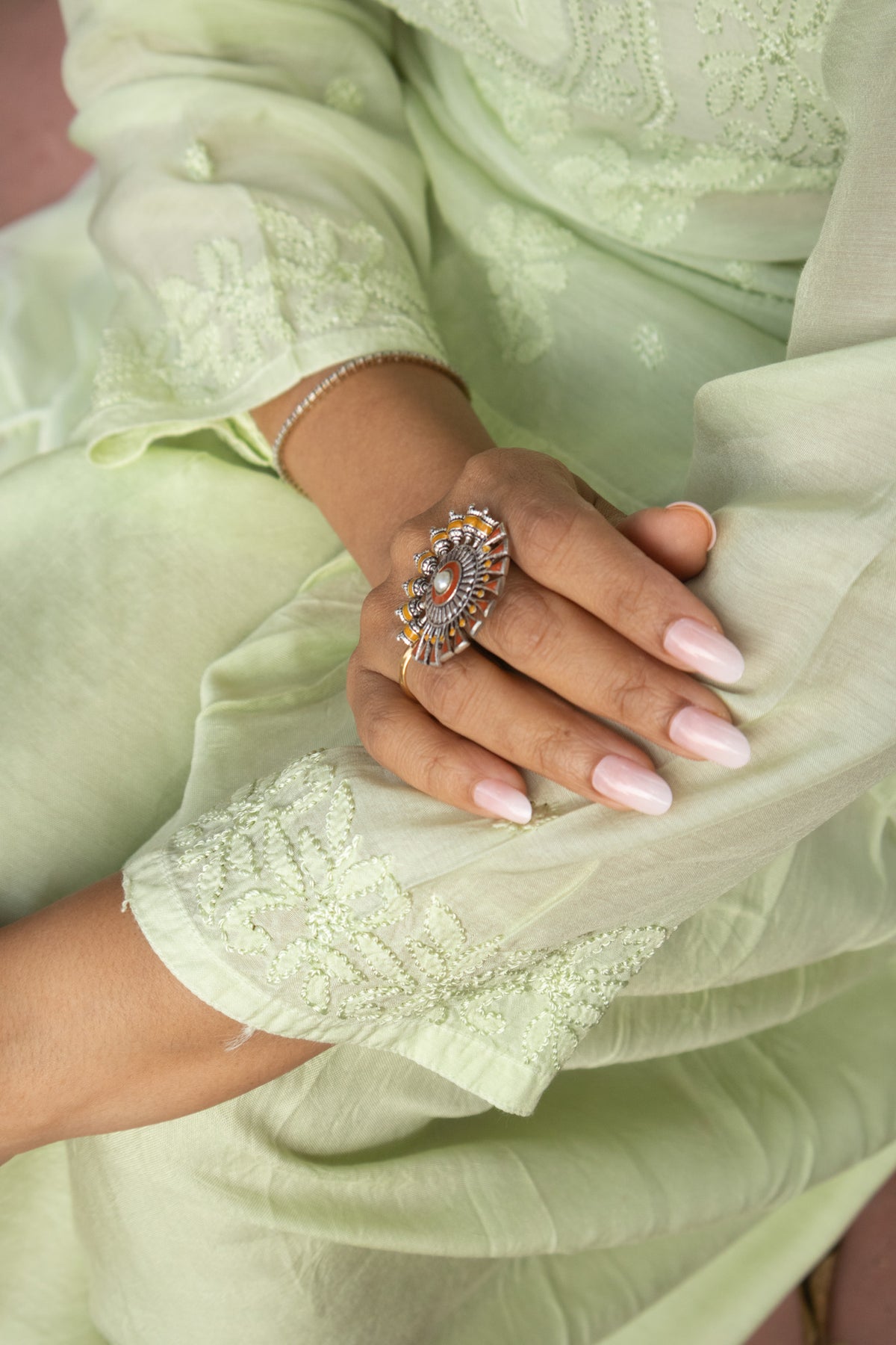 A close-up of a person's hand wearing a large, ornate gold ring with a central blue and white stone, styled with a light green embroidered traditional outfit.