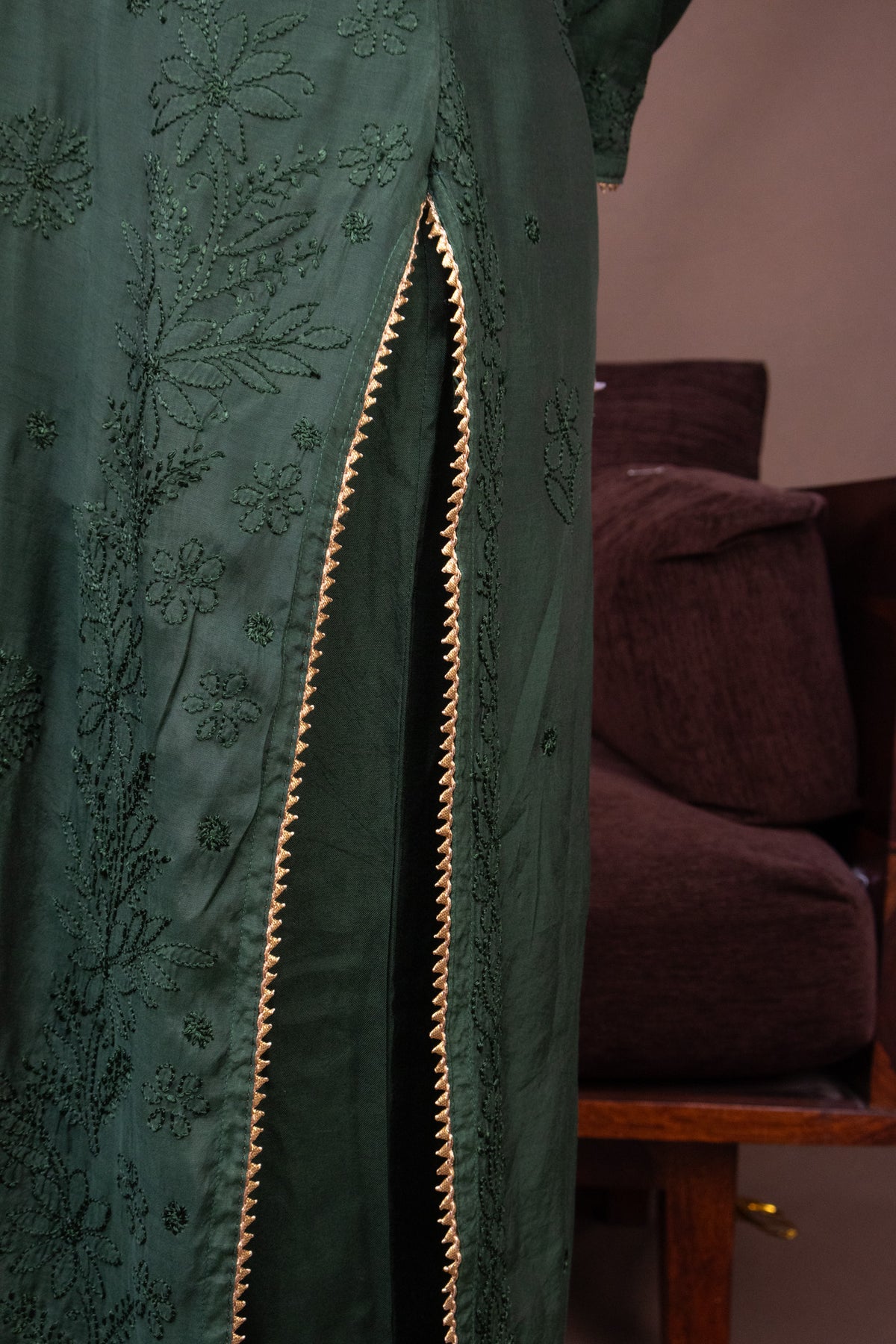 A close-up of a dark green embroidered robe or dress with a vertical slit outlined by a decorative gold braided trim, shown indoors next to a brown upholstered chair.