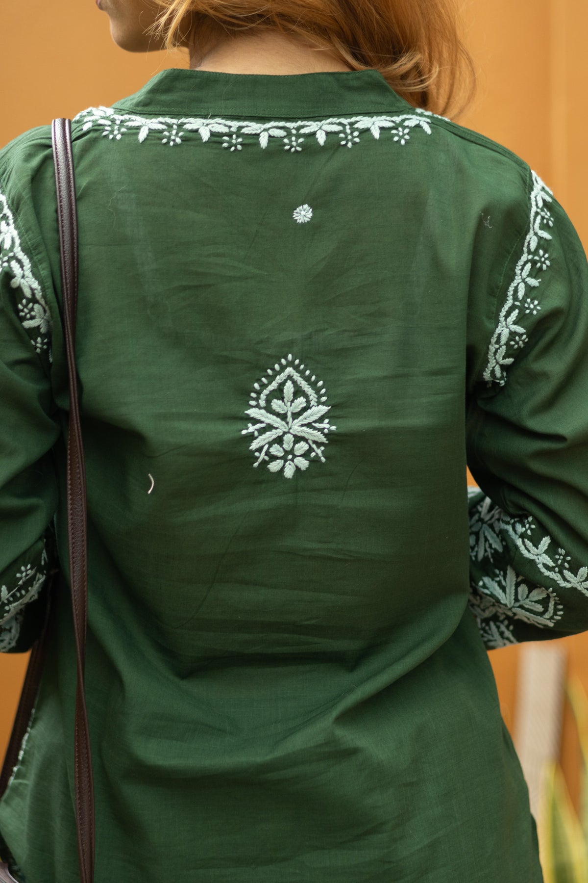 A close-up back view of a person wearing a dark green embroidered tunic or kurta. The garment features white floral and geometric embroidery along the neckline, sleeves, and a central motif on the back.