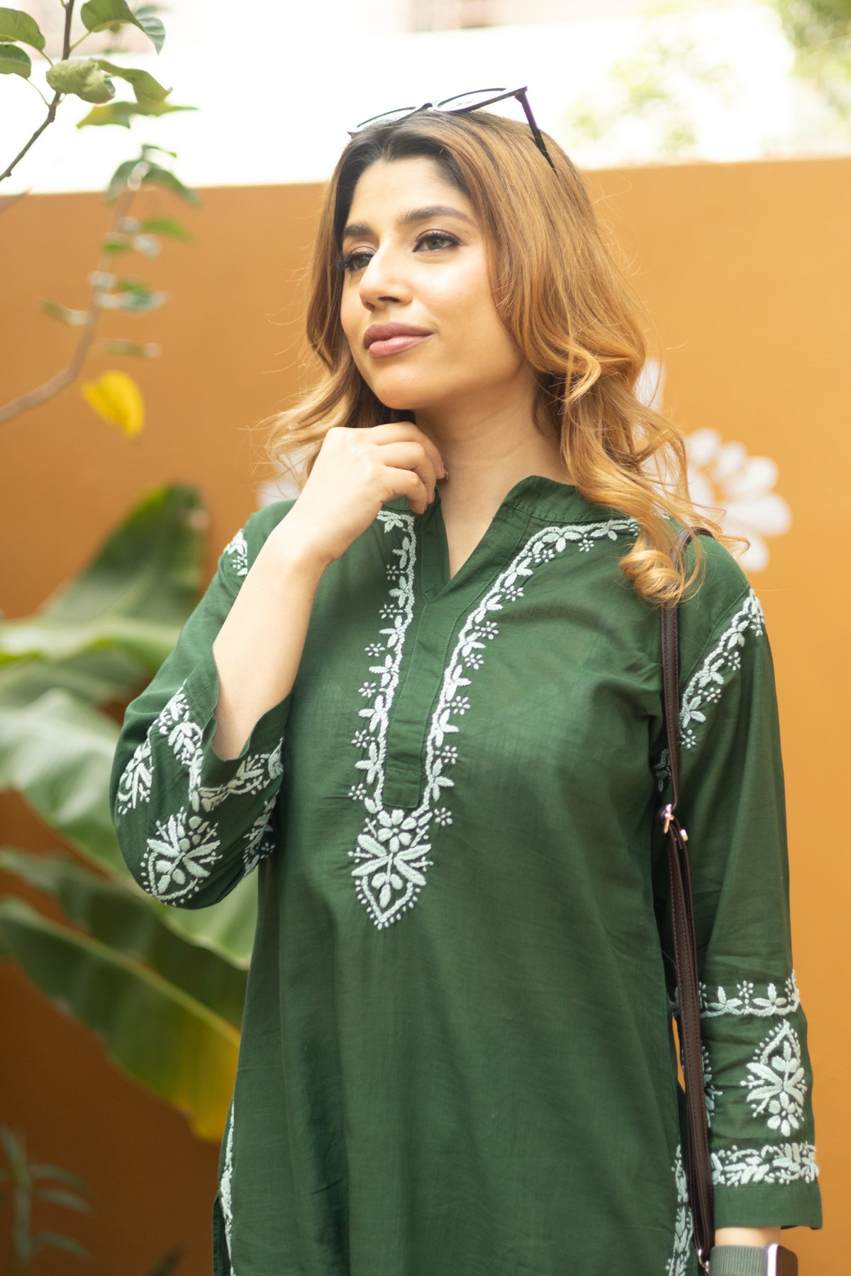 A woman wearing a dark green cotton tunic with white embroidery on the neckline and cuffs, standing against an orange wall with green plants in the background.