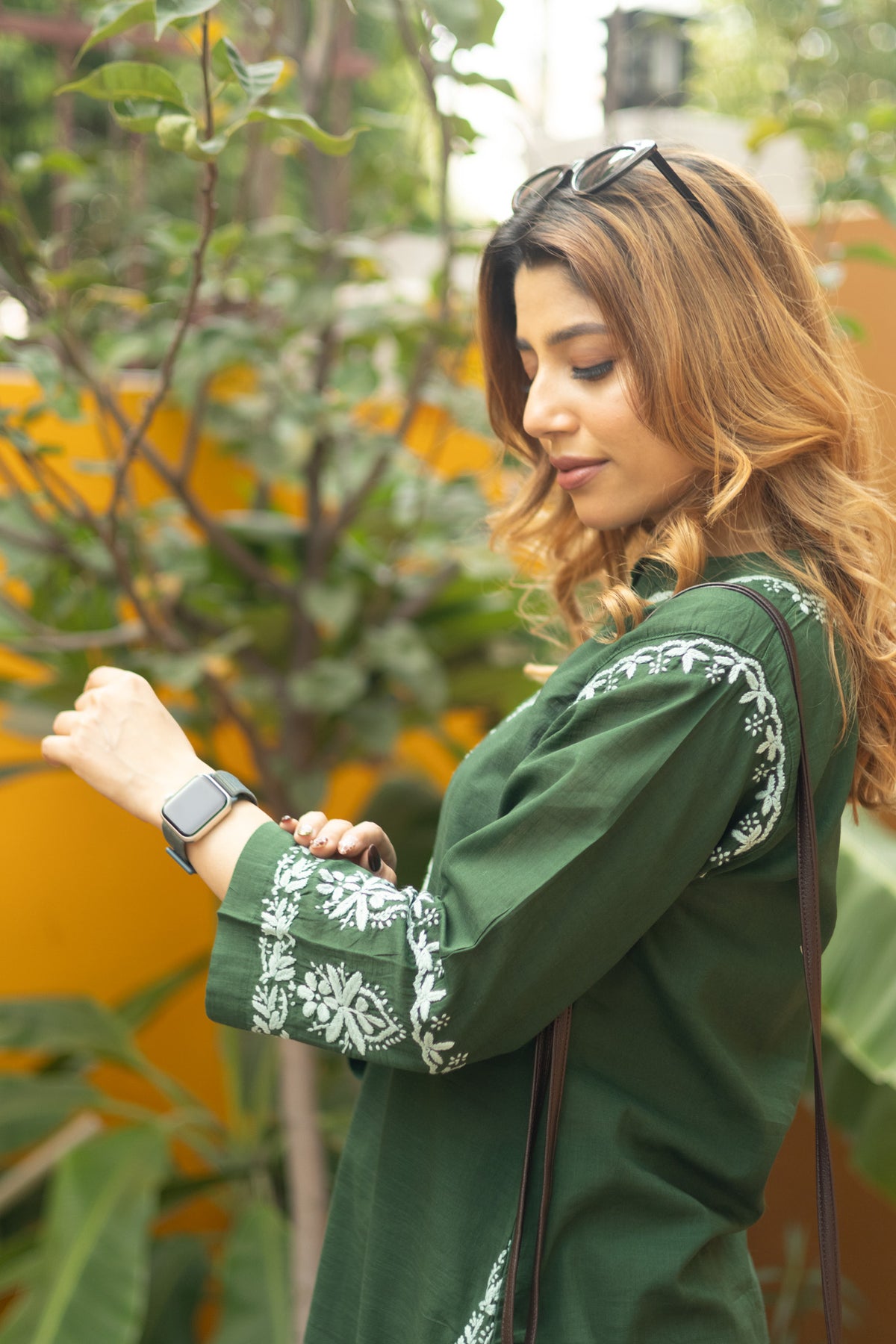A woman in a dark green embroidered tunic with white floral patterns on the cuffs, standing outdoors in front of a yellow wall and green plants.