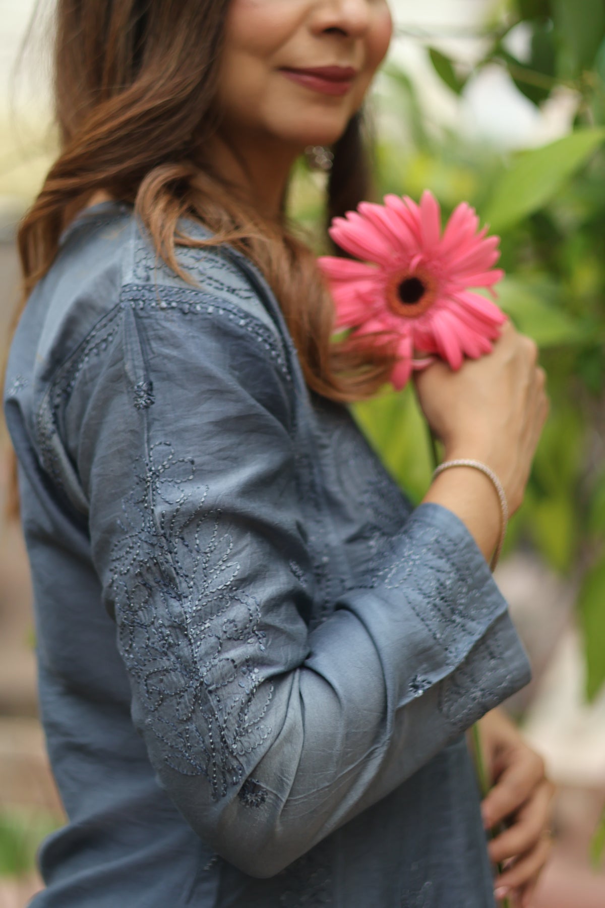 A woman wearing a dusty blue long-sleeve embroidered kurta, holding a pink flower against a soft-focus green garden background.