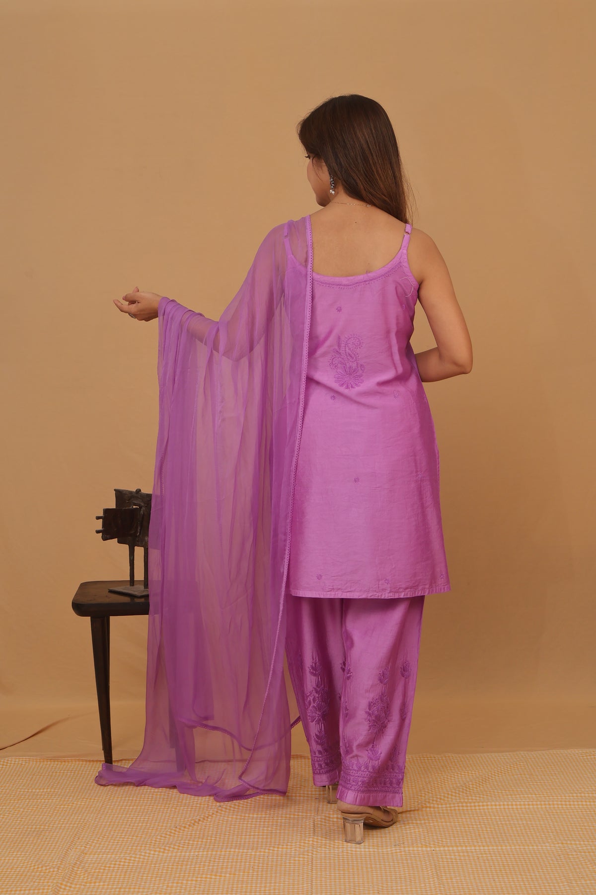 A woman wearing a purple three-piece traditional outfit with a matching sheer dupatta, standing against a plain beige background.