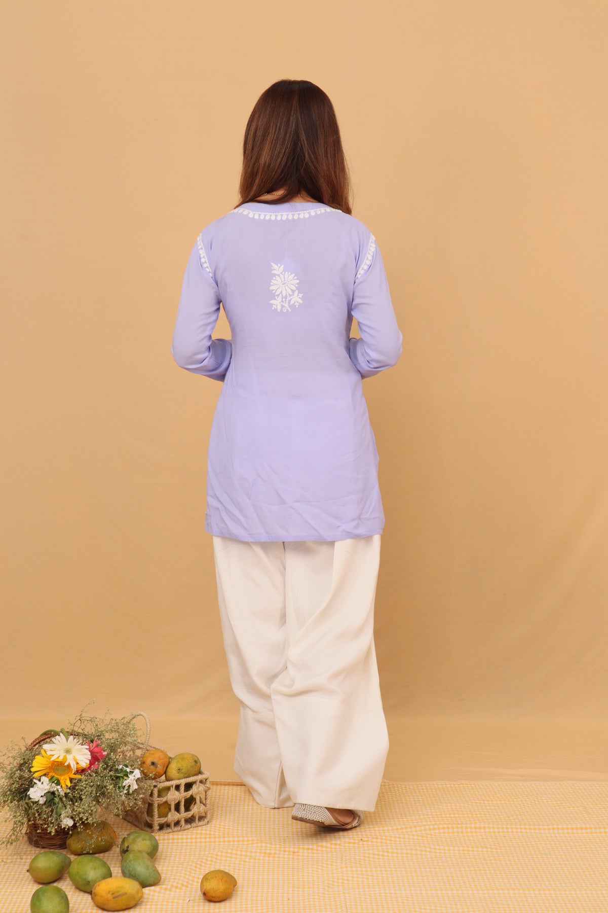 A woman stands with her back to the camera wearing a light lavender long-sleeve kurta with white embroidery on the back and white wide-leg pants, set against a solid beige backdrop with decorative fruit and flowers on the floor.