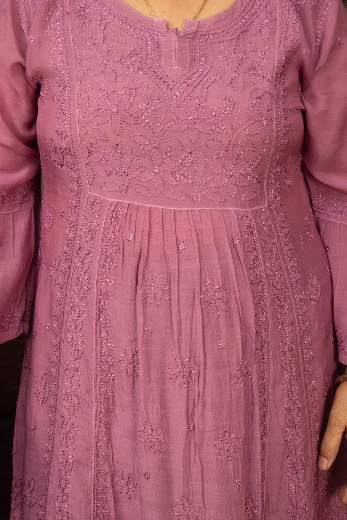 A close-up of a dusty pink or mauve-colored embroidered tunic or dress, showing detailed floral embroidery and beadwork on the bodice and pleated skirt.