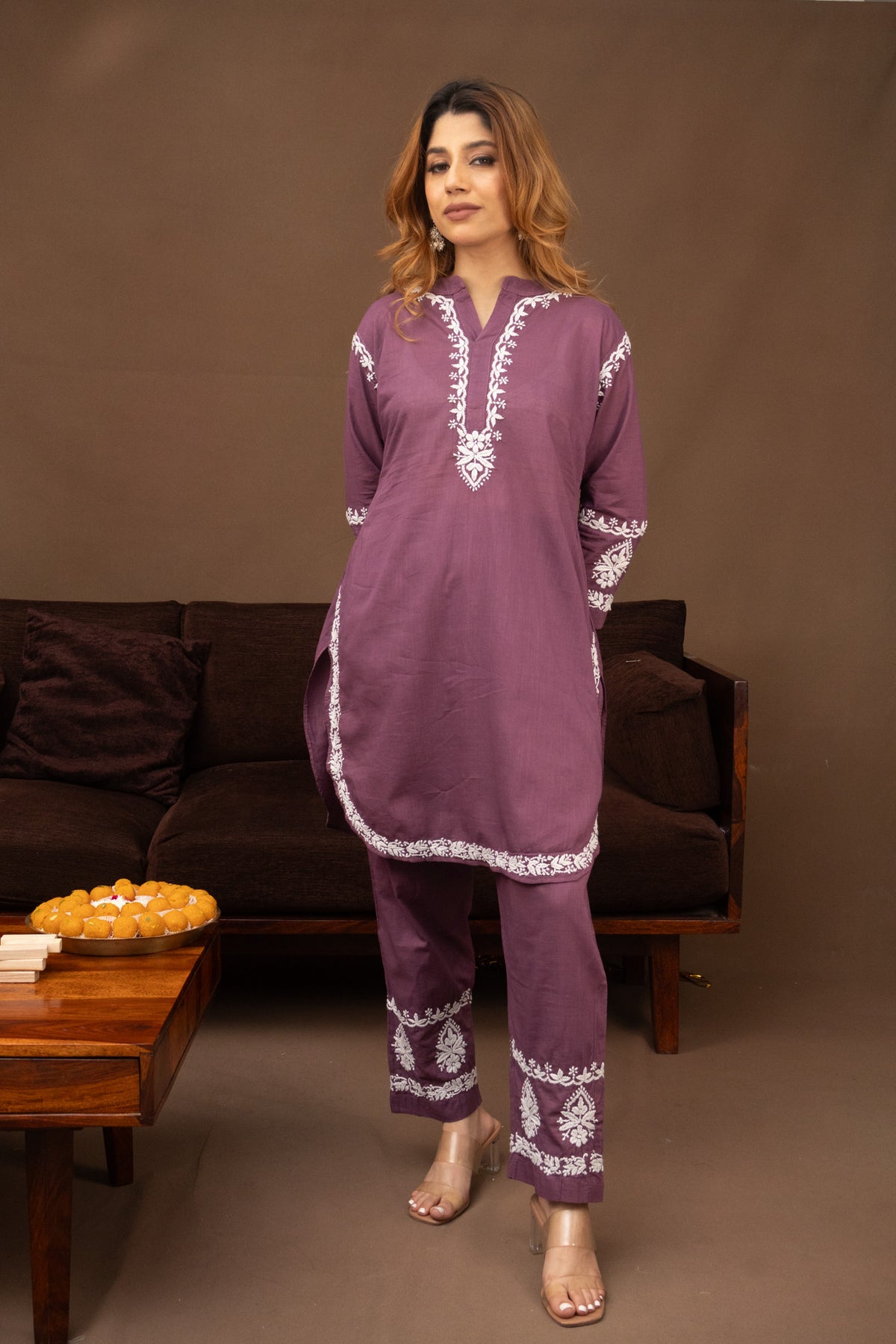 A woman modeling a two-piece purple kurta and trouser set with white embroidery, standing in front of a brown sofa in a living room setting.