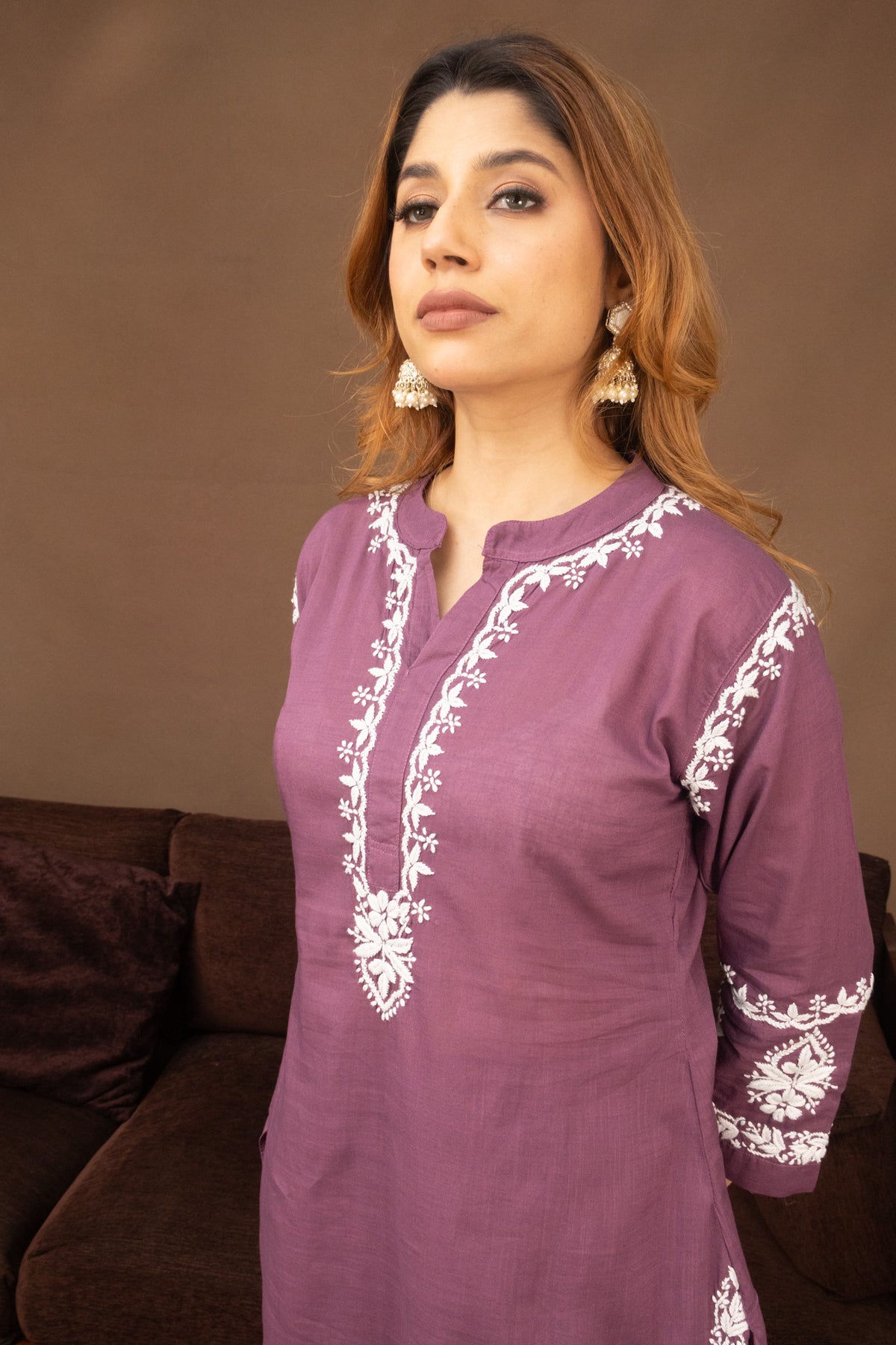 A woman wearing a mauve purple kurta with white floral embroidery on the neckline, sleeves, and hem, standing in front of a brown background.