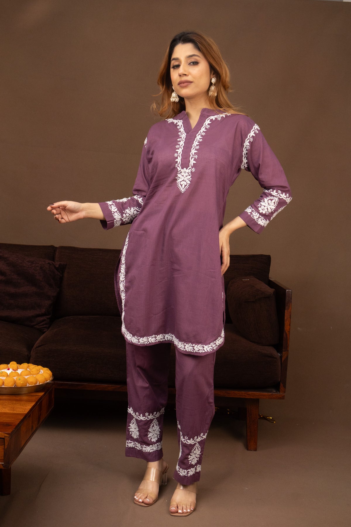 A woman models a purple two-piece kurta and trouser set with white embroidery, standing in front of a brown sofa.