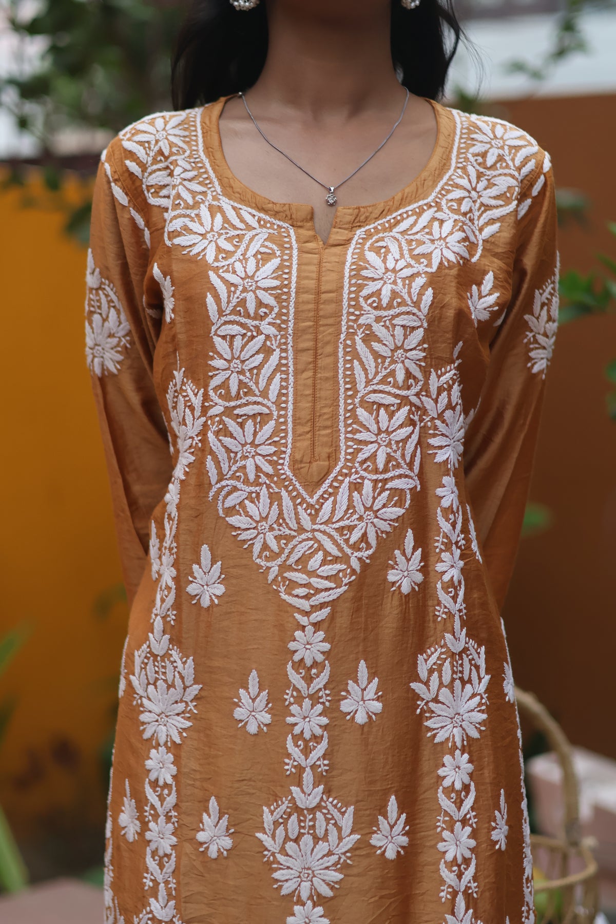 A woman wearing a mustard yellow long-sleeve tunic with intricate white floral embroidery, photographed from the neck down against an outdoor background.