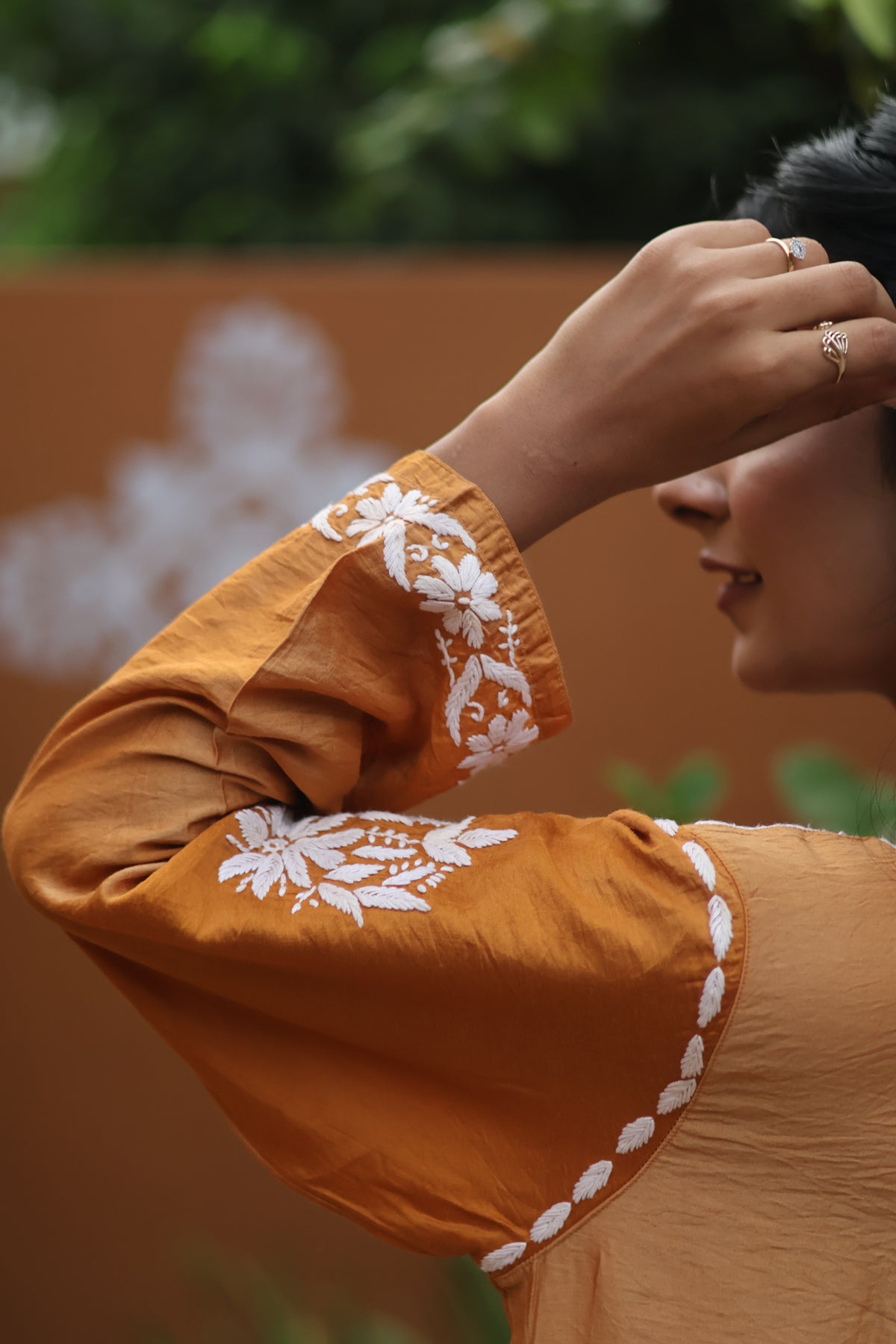 A close-up lifestyle shot of a woman wearing a mustard yellow kurta with white floral embroidery on the sleeve cuffs, set against a warm brown wall and green foliage background.