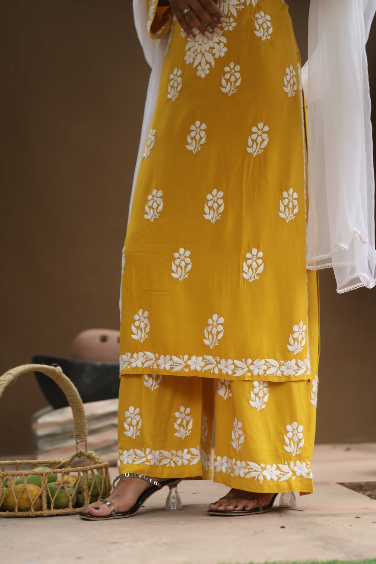 A person wearing a mustard yellow and white printed traditional outfit with wide-leg pants and a white draped overlay, shown from the waist down with clear-strap heels. A woven basket and stacked books are visible against a warm brown background.