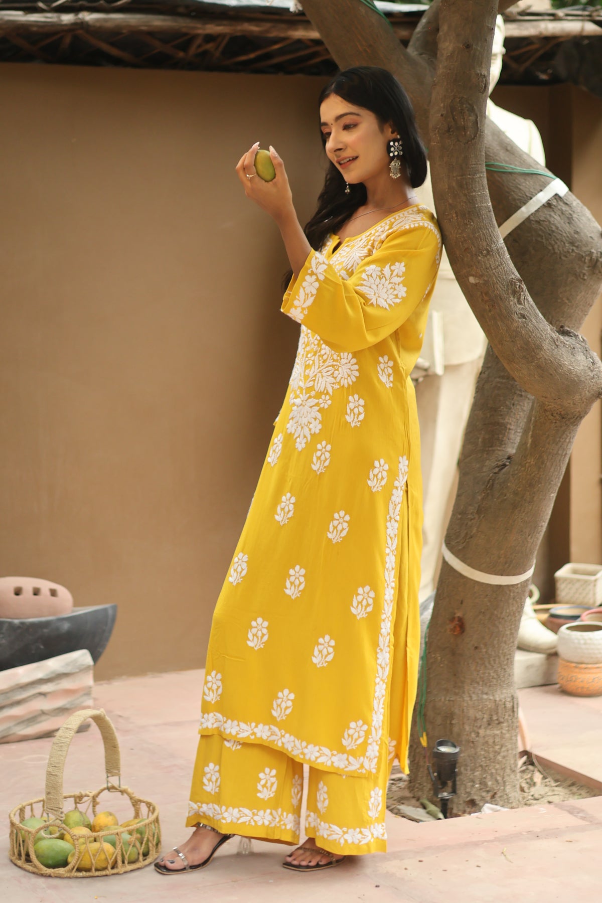 A woman wearing a bright yellow traditional dress with white floral embroidery and border details, standing in an outdoor setting near a tree trunk.