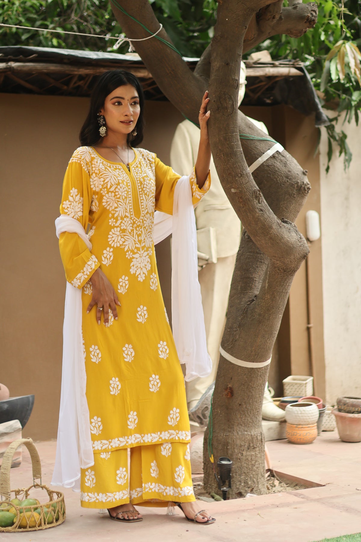 A woman models a yellow and white embroidered Indian salwar kameez suit in an outdoor courtyard setting next to a tree.