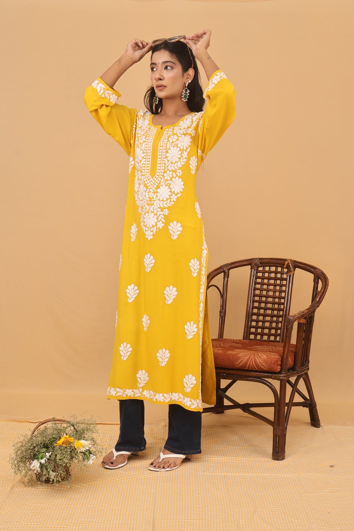 A woman models a bright yellow traditional kurta with white floral embroidery and white border trim, paired with black pants, standing next to a wooden chair against a beige background.
