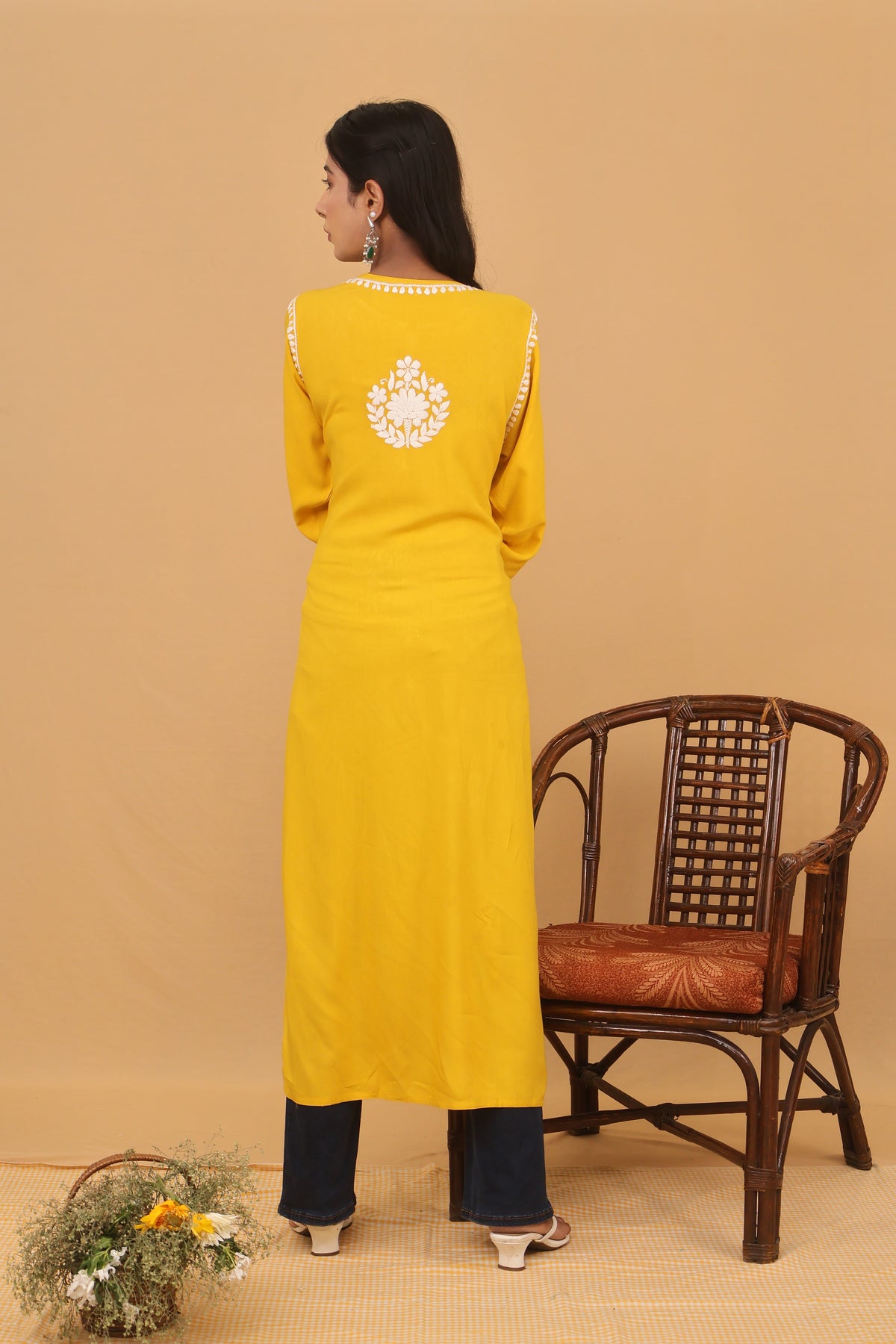 A woman wearing a bright yellow traditional outfit with a white embroidered circular motif on the back and dark navy pants, standing next to a brown wooden chair against a beige background.