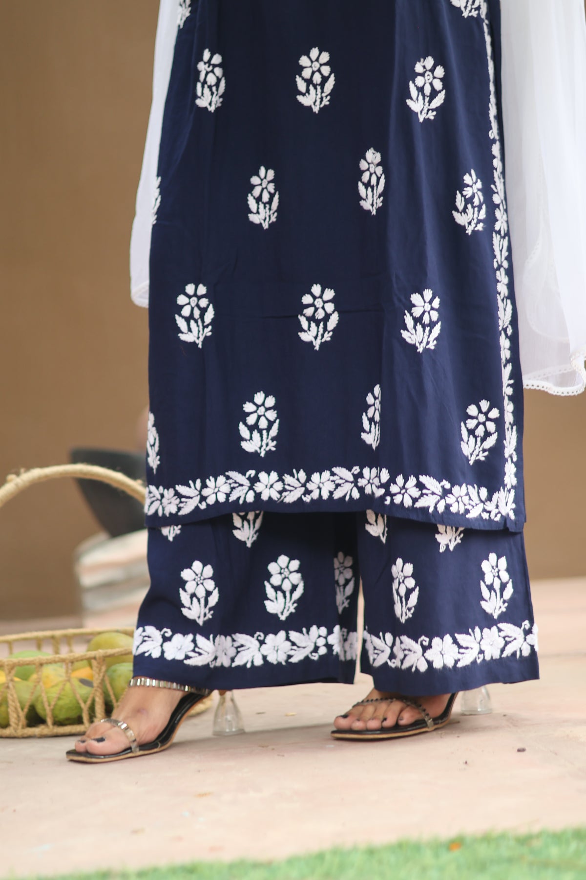 A close-up of a person wearing a navy blue traditional outfit with white floral block prints and matching wide-leg pants, paired with clear high-heeled sandals.