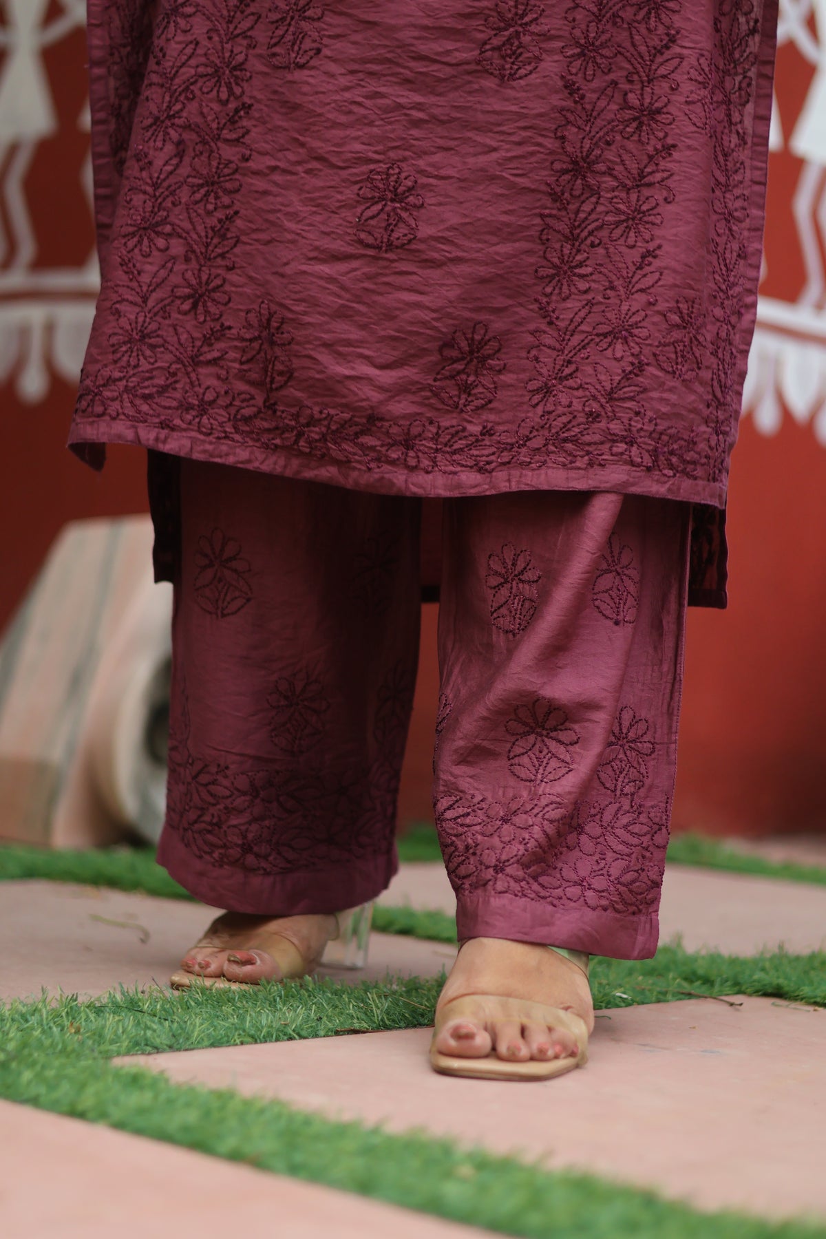 A close-up of a person's feet wearing a traditional maroon salwar kameez set with floral embroidery, standing on a tiled surface with artificial grass.