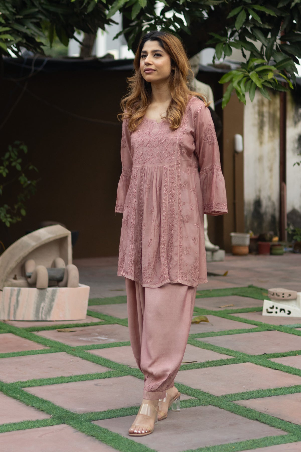 A woman modeling a dusty rose pink ethnic kurta and pant set in an outdoor courtyard setting with green grass accents in the paving.