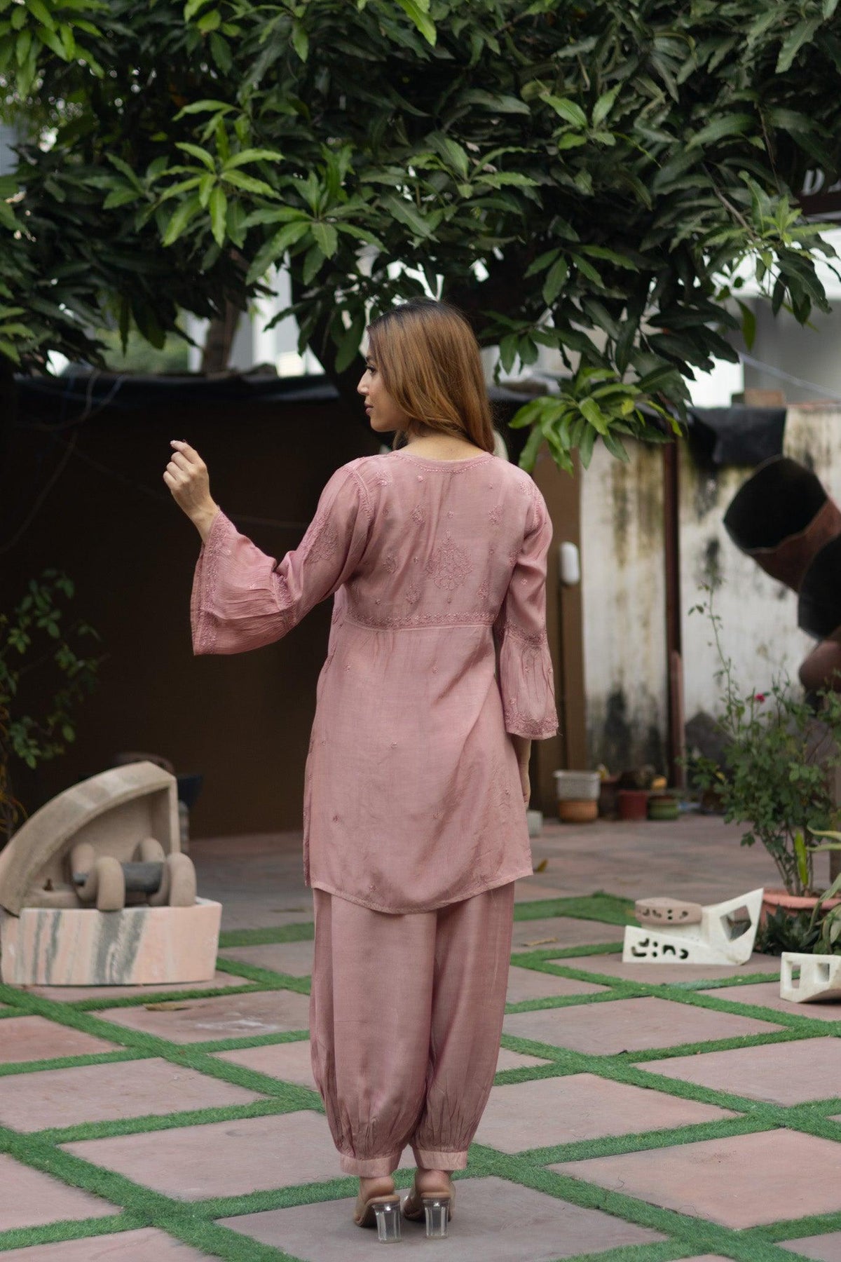 A woman wearing a dusty pink two-piece tunic and trouser set, photographed from behind in an outdoor courtyard setting with greenery and potted plants. Onion Muslin Nyra Cut Chikankari Kurti with Bakhiya Embroidery