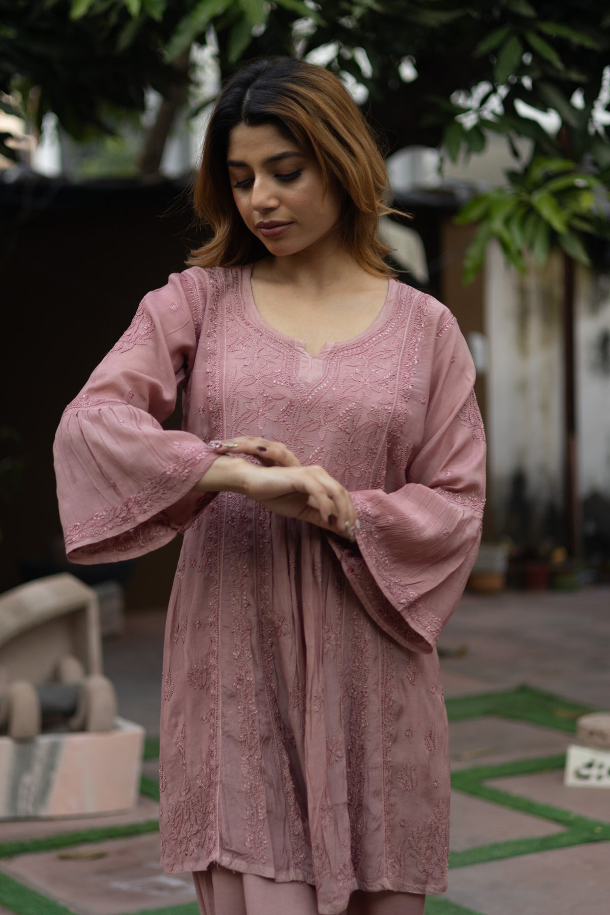 A woman wearing a dusty pink embroidered tunic with wide bell sleeves, standing outdoors in a garden courtyard.