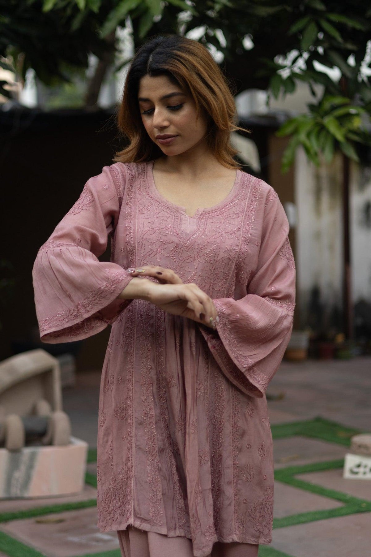 A woman wearing a dusty pink embroidered tunic with wide bell sleeves, standing outdoors in a garden courtyard. Onion Muslin Nyra Cut Chikankari Kurti with Bakhiya Embroidery