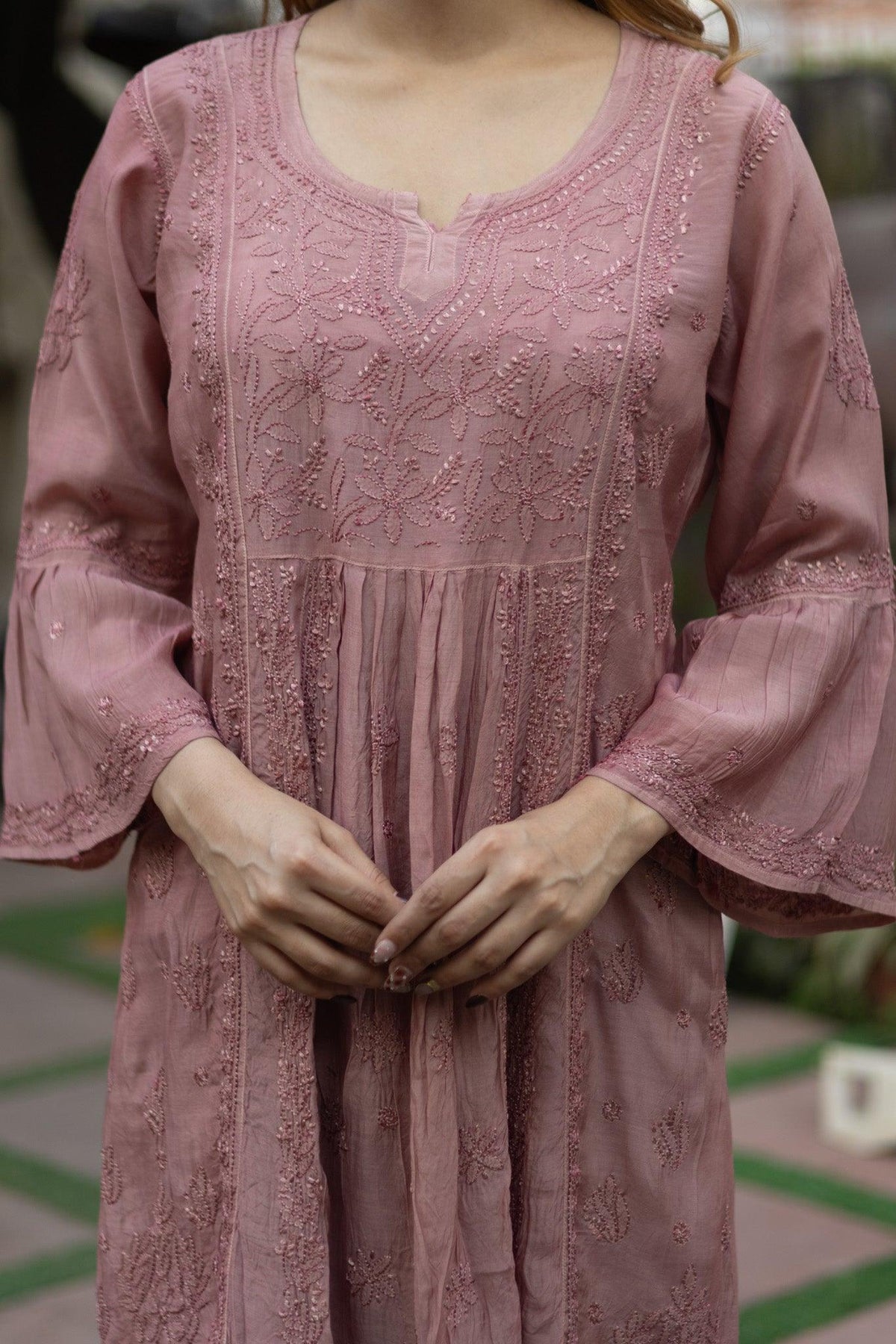 A dusty pink embroidered tunic dress with long bell sleeves and a V-neckline, shown on a model with hands clasped in front. Onion Muslin Nyra Cut Chikankari Kurti with Bakhiya Embroidery
