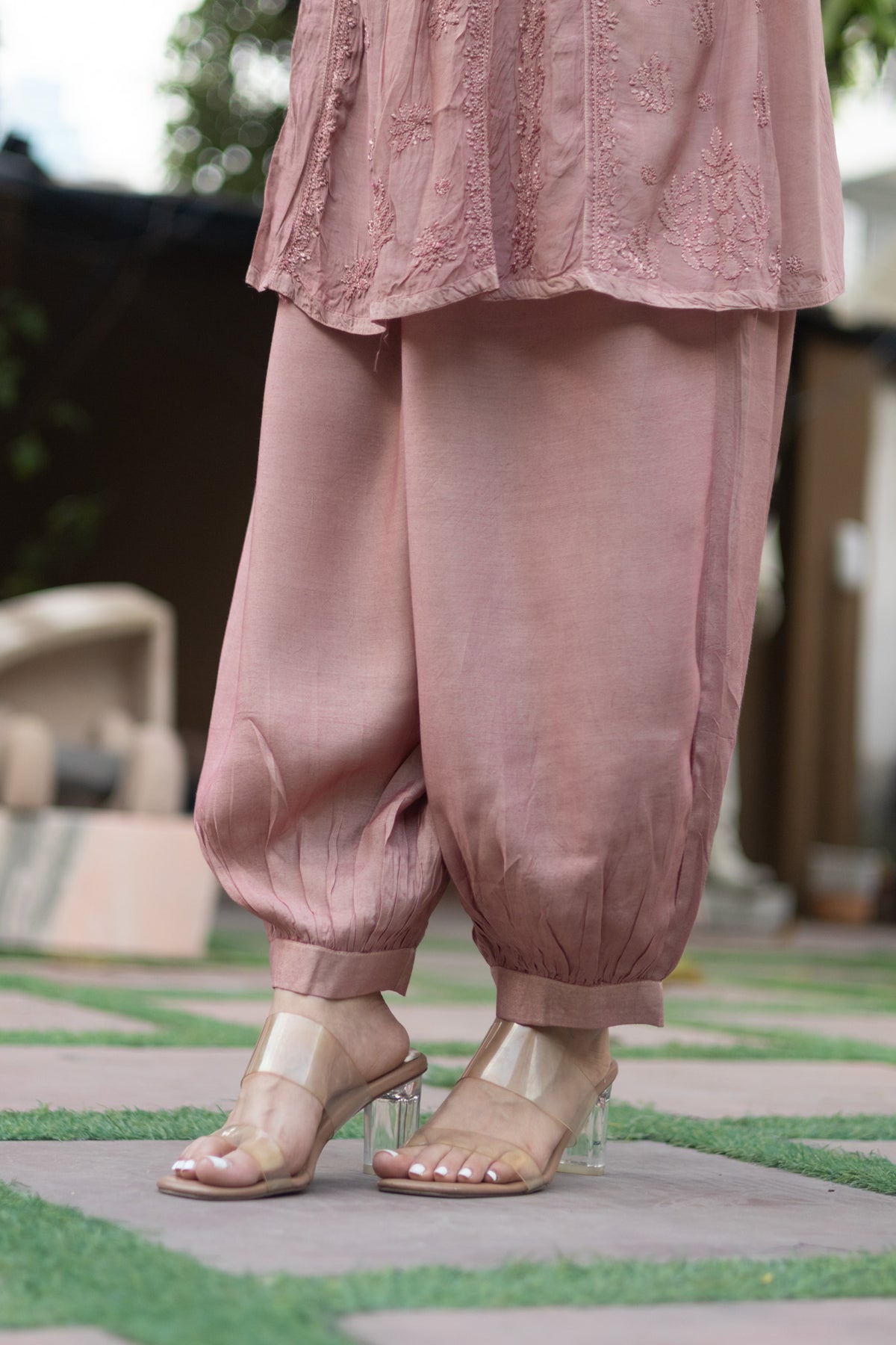 A person wearing dusty pink harem-style pants and a matching embroidered top, paired with clear high-heel sandals, standing outdoors on a paved path with green grass.