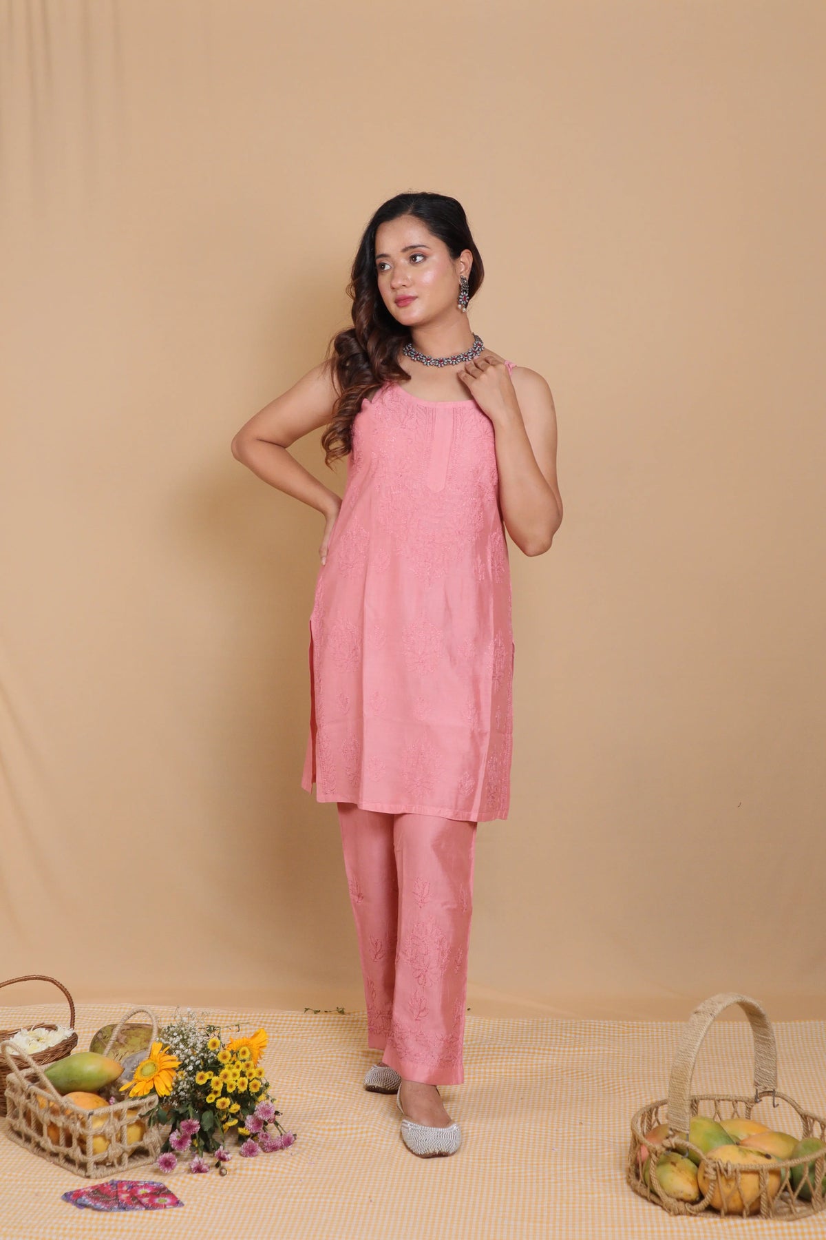 A woman modeling a pink sleeveless kurta and matching trousers set, standing against a beige backdrop with decorative baskets and flowers on the floor.