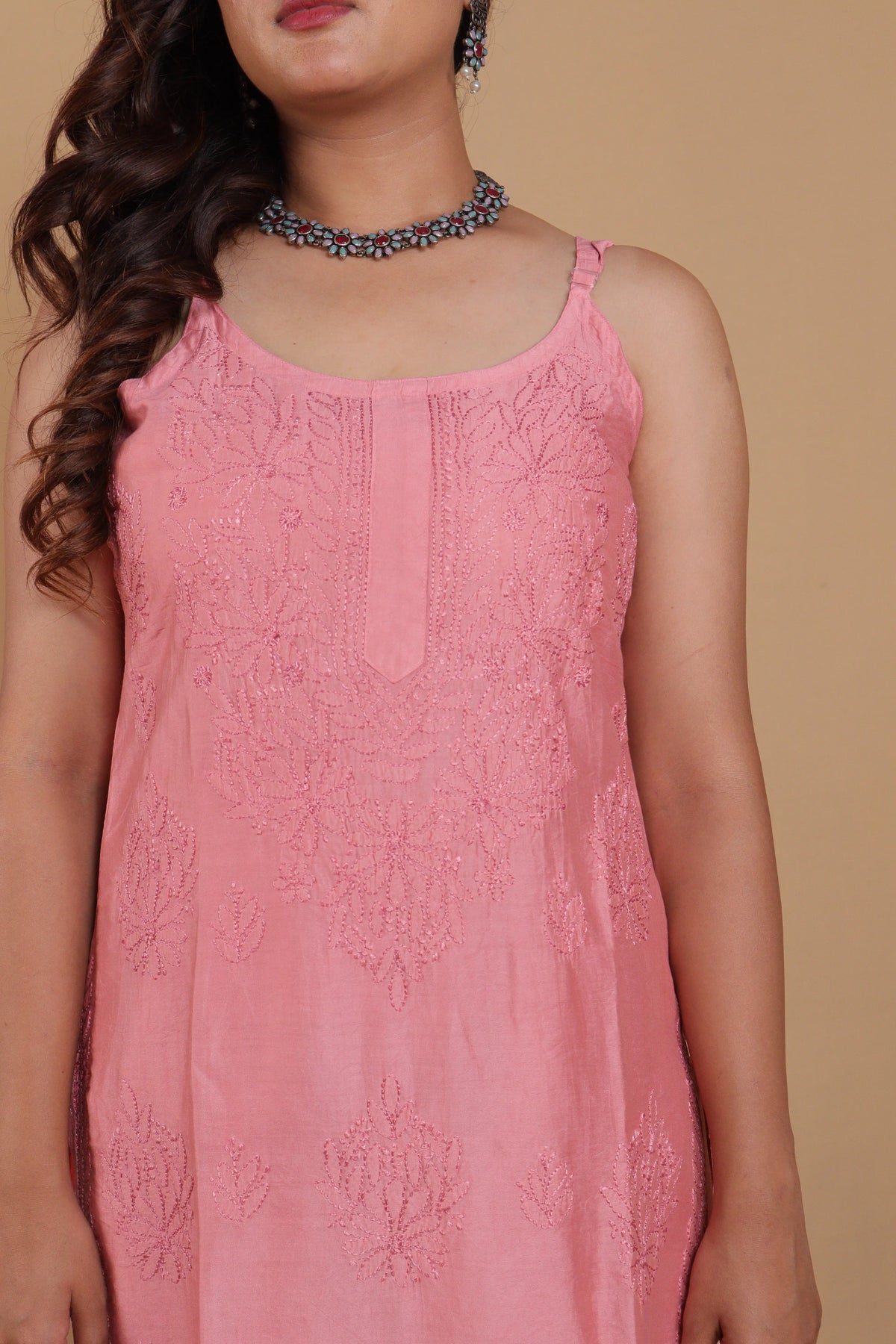 A woman wearing a pink sleeveless embroidered dress or tunic with floral lace detailing, shown against a plain beige background.
