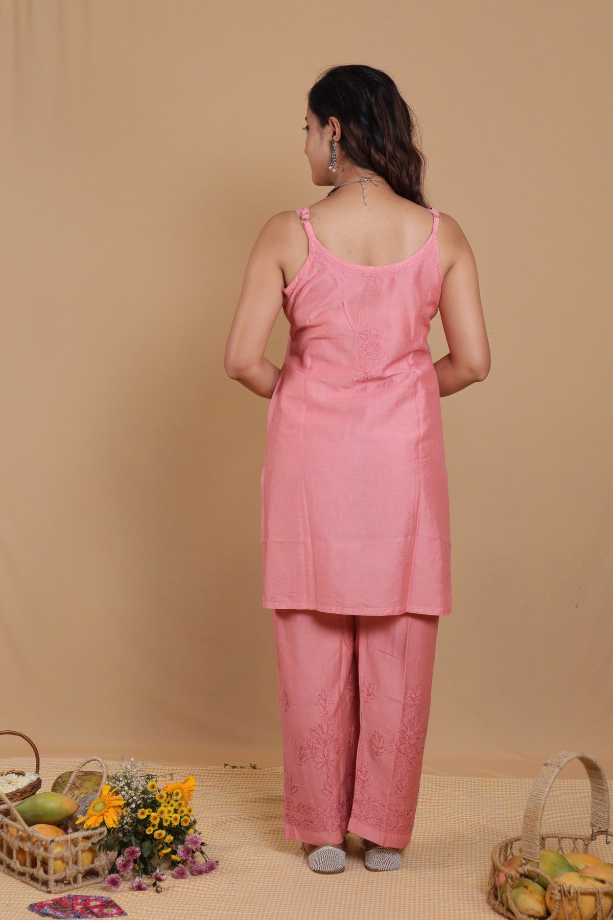 A woman stands with her back to the camera wearing a light pink two-piece kurta set with a satin-like sleeveless top and matching wide-leg trousers. The set is photographed against a plain beige backdrop with decorative baskets and flowers on the floor.