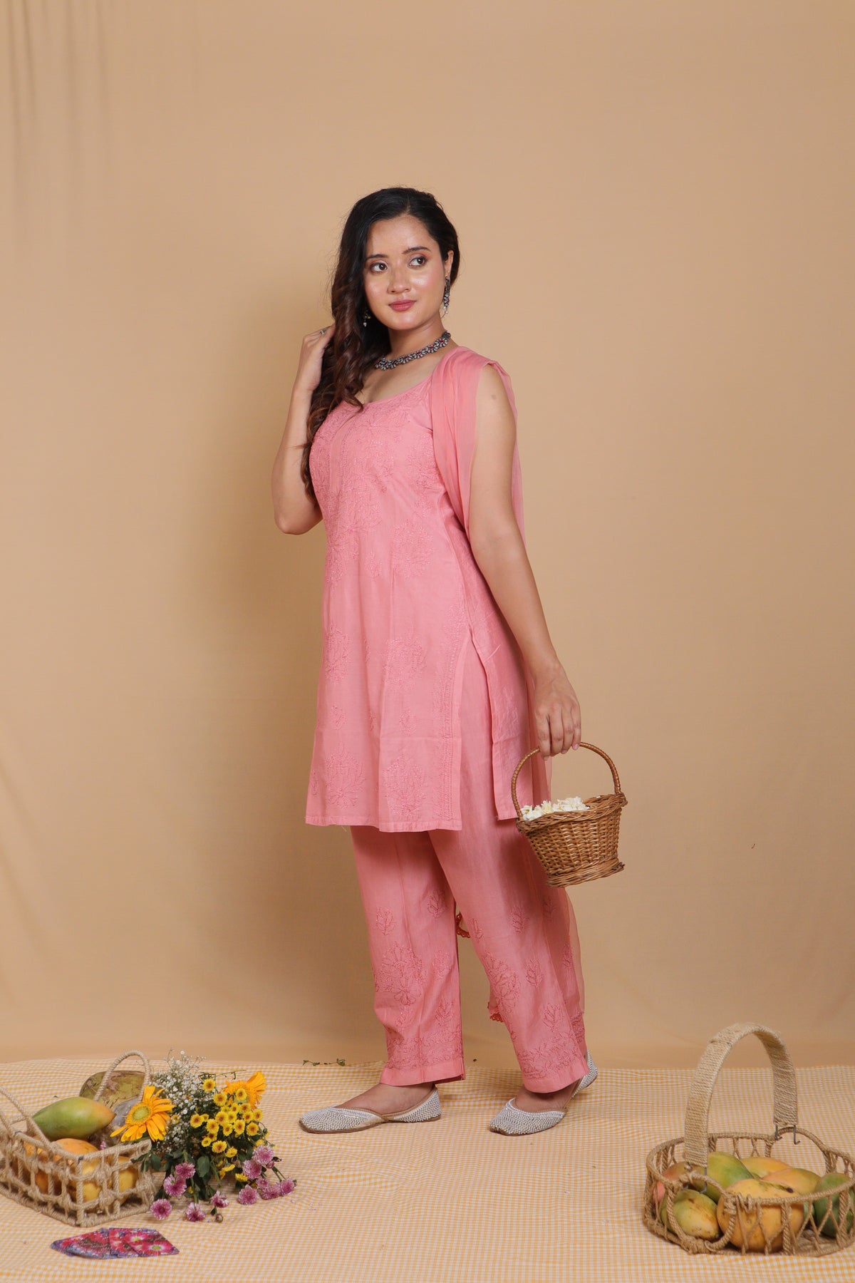 A woman modeling a pink two-piece kurta set with matching pants, standing against a beige backdrop and holding a small woven basket.