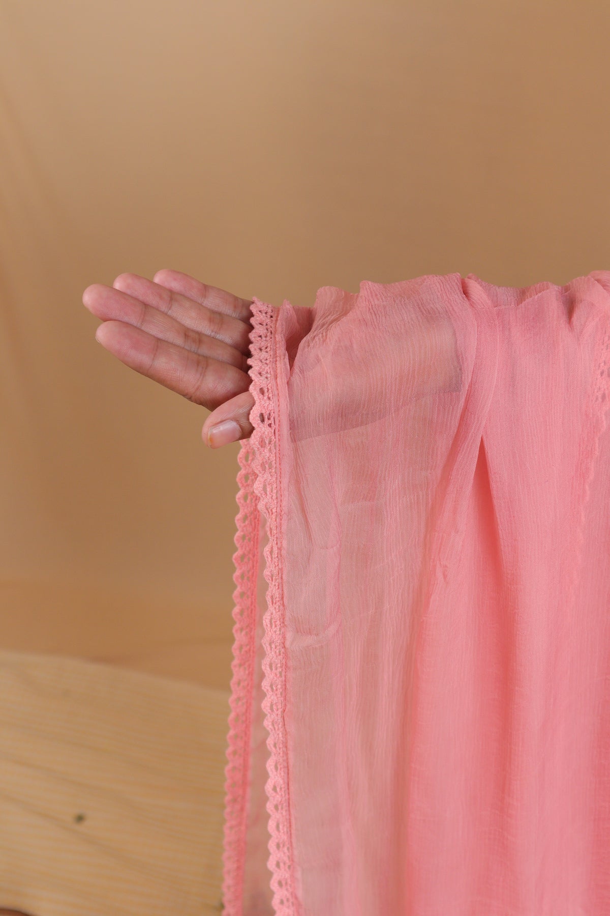A close-up of a hand holding up a sheer pink fabric, showing its texture and a decorative scalloped lace trim, against a warm beige background.