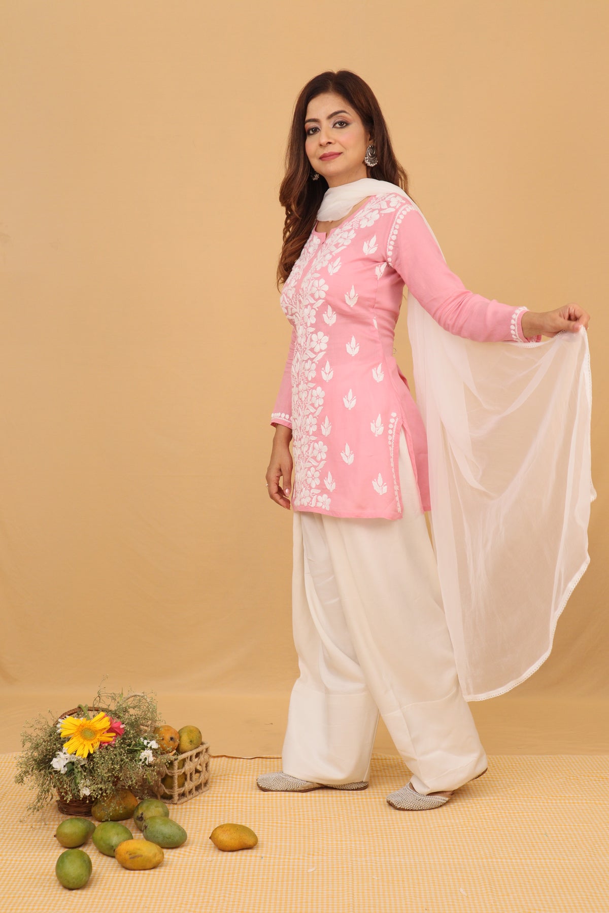 A woman modeling a traditional Indian outfit consisting of a pink kurta with white floral embroidery, white palazzo pants, and a white dupatta, posed against a solid beige studio backdrop with decorative fruits and flowers on the floor.