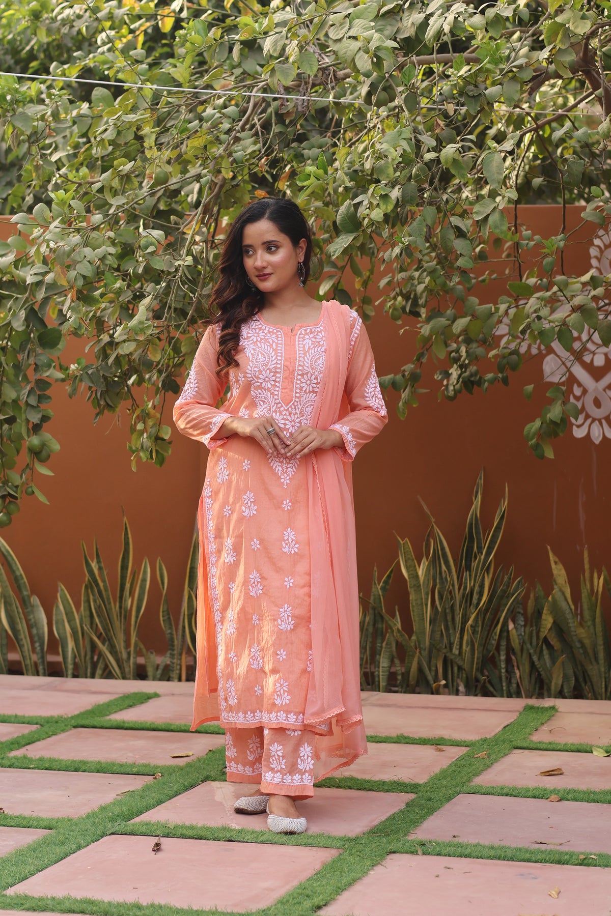 A woman wearing a peach-colored embroidered kurta set with matching pants and a dupatta, standing outdoors on a tiled patio with green plants and a terracotta wall behind her.
