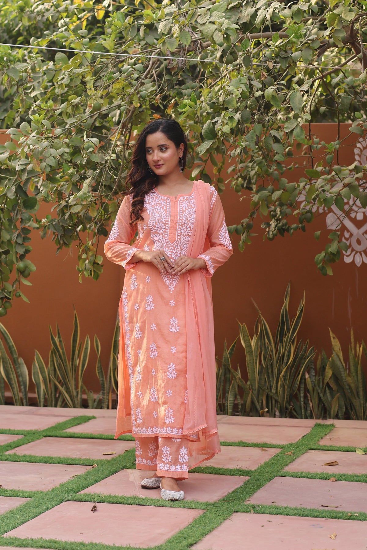 A woman wearing a peach-colored embroidered kurta set with matching pants and a dupatta, standing outdoors on a tiled patio with green plants and a terracotta wall behind her. Peach Mul Chanderi White Thread Chikankari Ghaspatti 3 Piece Set with Dupatta