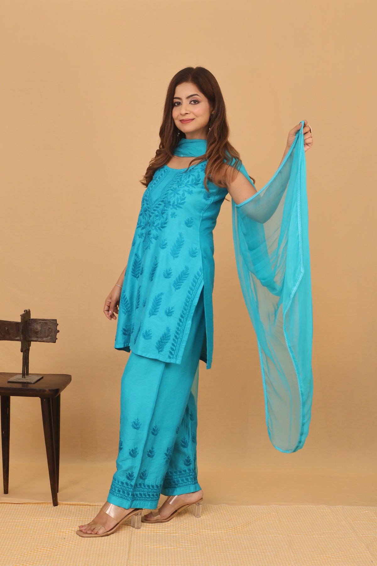 A woman models a turquoise blue ethnic salwar kameez suit with matching dupatta against a solid beige backdrop. The set features a printed kurta, matching trousers, and a sheer dupatta.