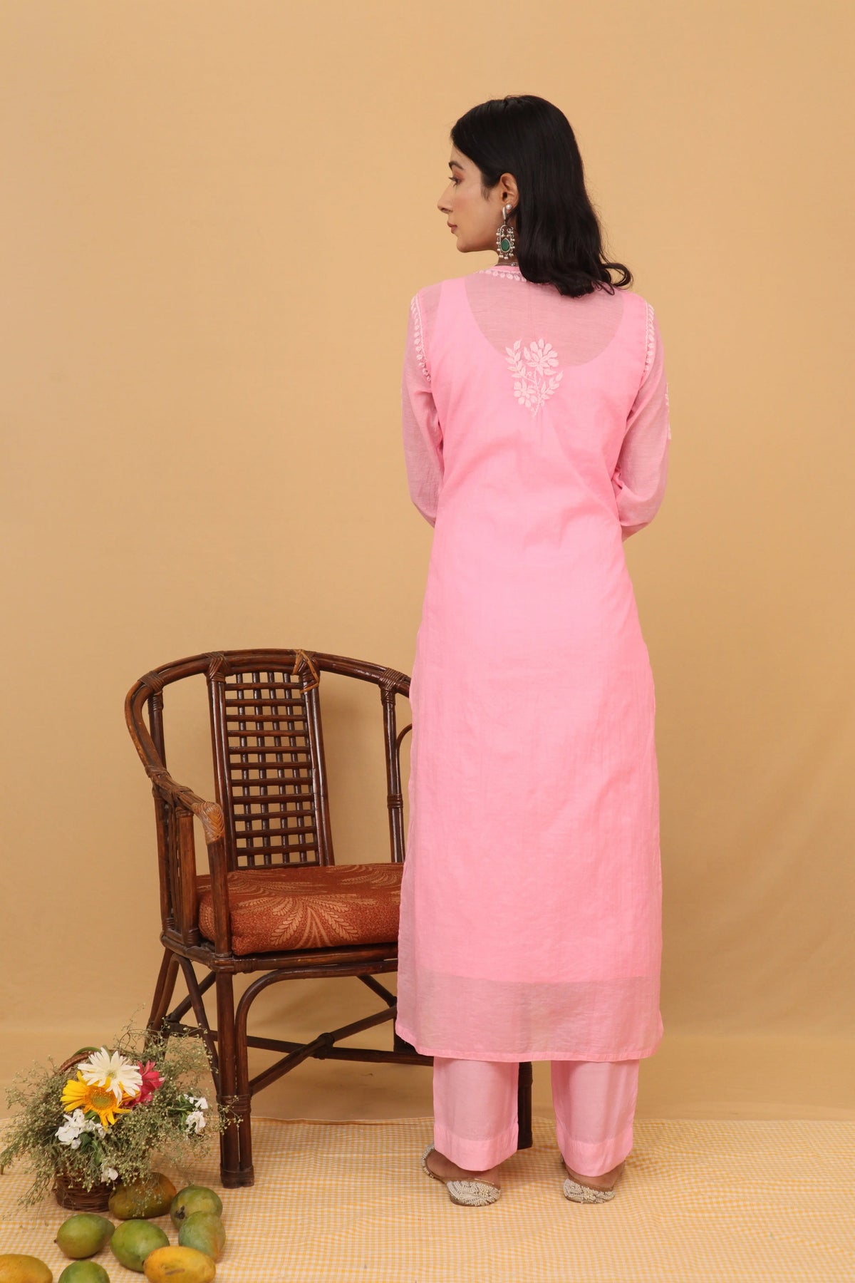 A woman stands with her back to the camera wearing a light pink traditional kurta and matching trousers. The outfit features white embroidered detailing on the upper back and sleeve cuffs.
