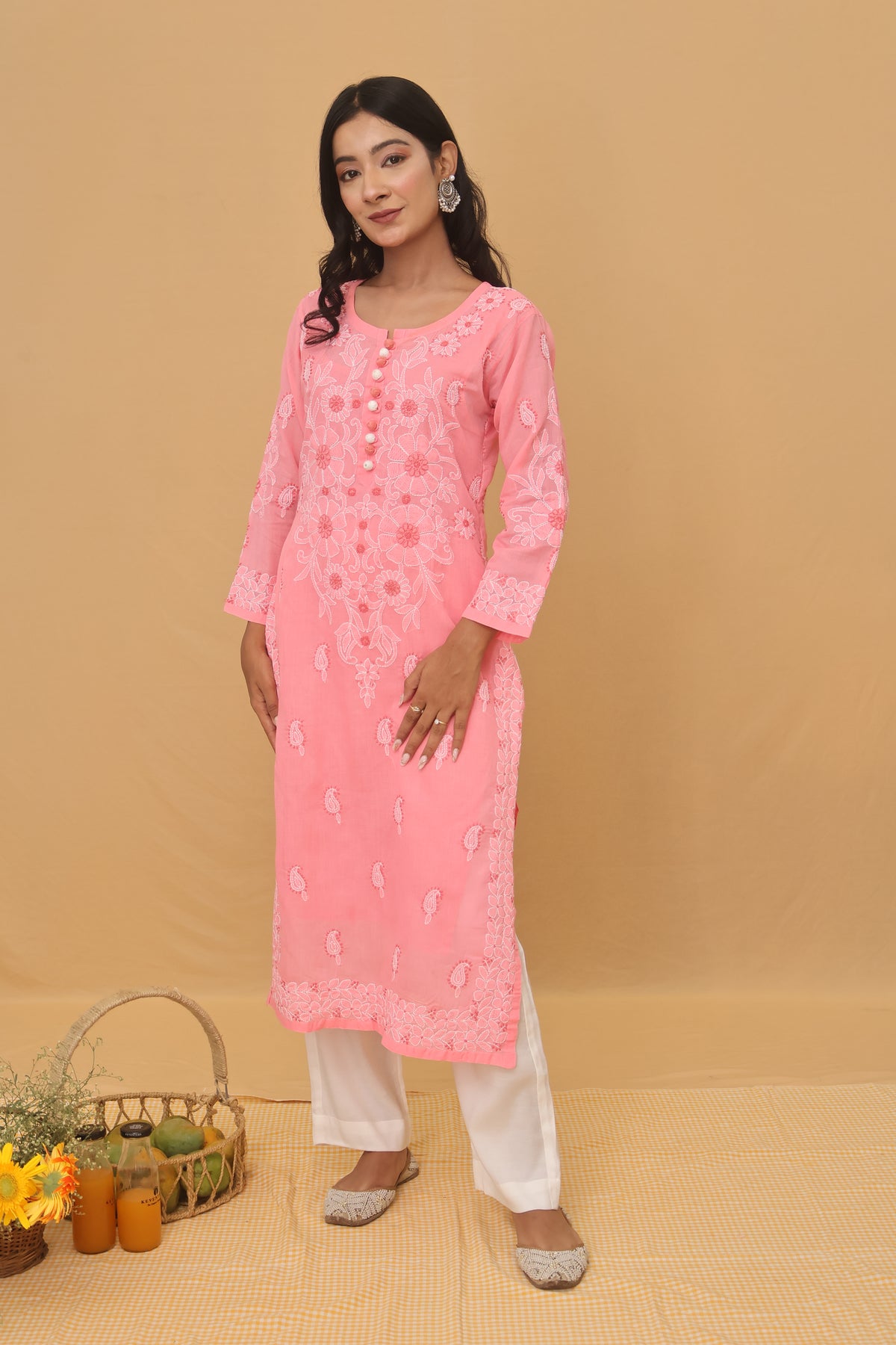 A woman modeling a pink embroidered kurta with white pants, standing against a solid beige backdrop. The kurta features light pink and white floral embroidery, and she is wearing silver flat shoes.