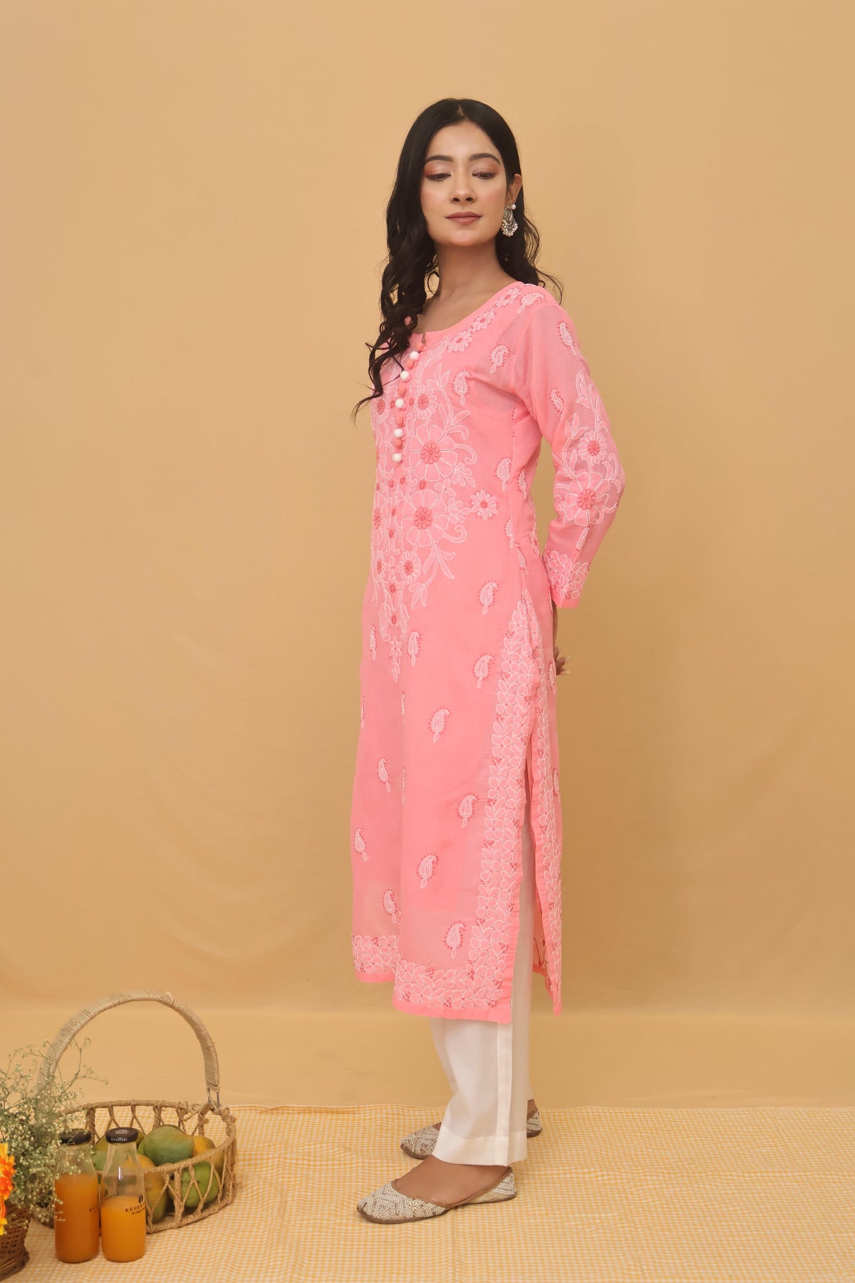A woman modeling a light pink embroidered kurta with white pants, standing against a solid beige backdrop. The kurta features delicate floral embroidery and a buttoned neckline.
