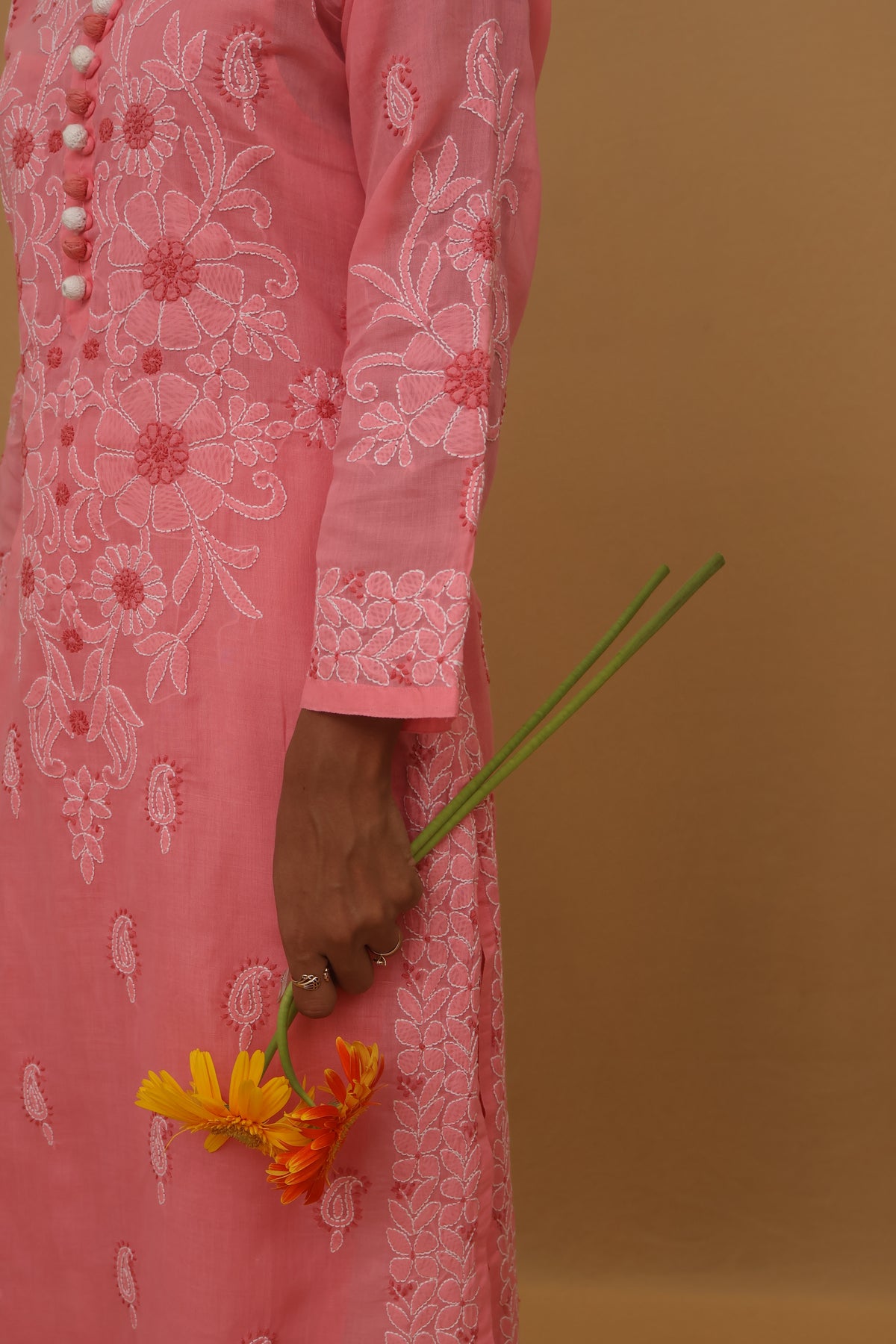 A close-up side view of a person wearing a pink embroidered traditional dress, holding a small yellow and orange flower against a solid warm brown background.