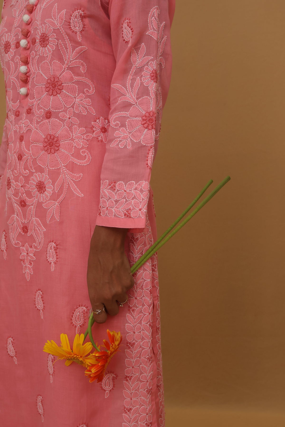 A close-up side view of a person wearing a pink embroidered traditional dress, holding a small yellow and orange flower against a solid warm brown background.