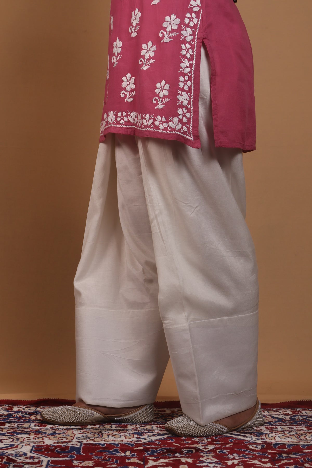 A person wearing a traditional outfit with a dusty pink embroidered kurta and off-white patola pants, standing on a patterned rug against a solid tan background.